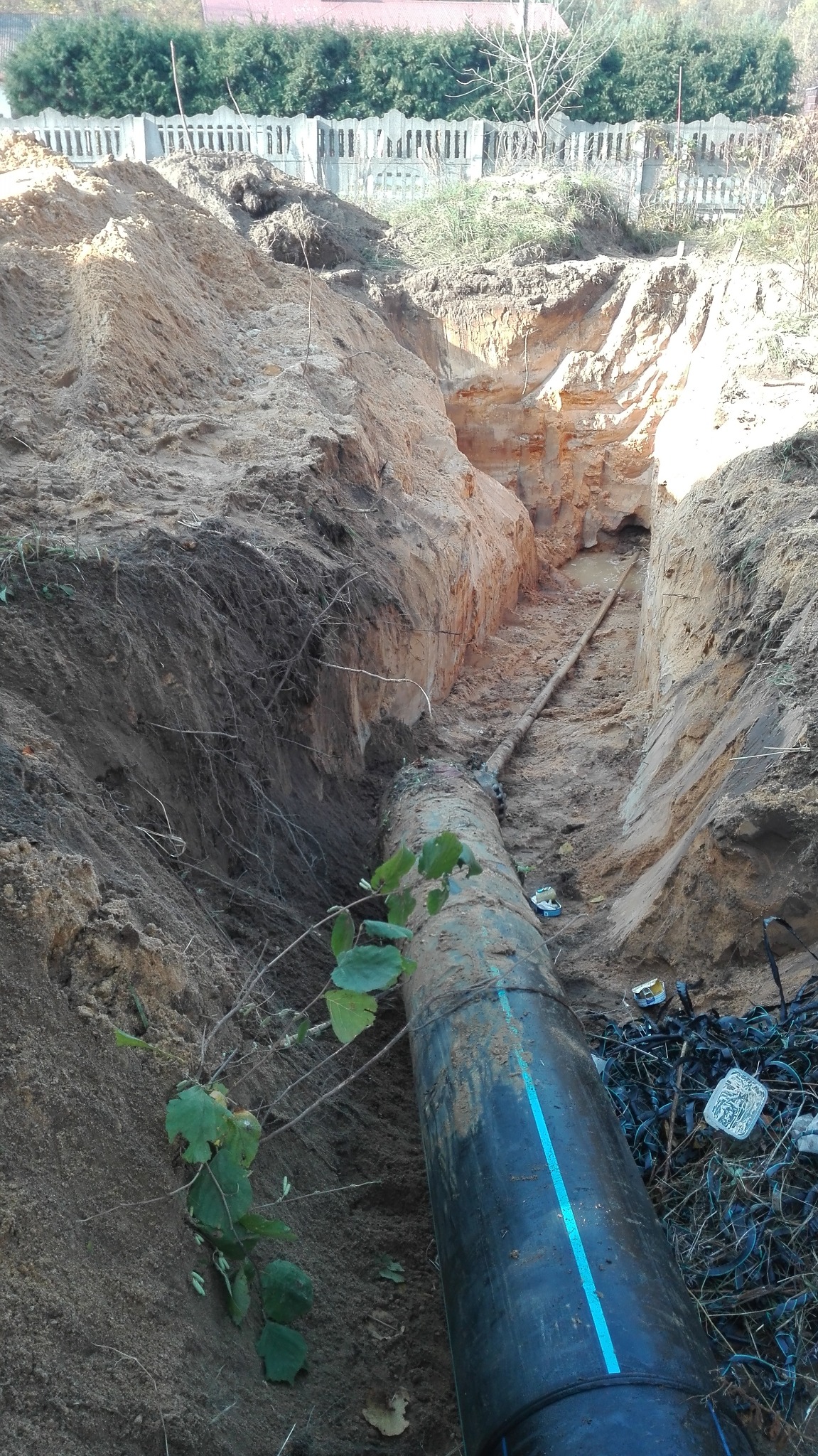Rura z niebieskim paskiem ułożona w wykopie ziemnym, widoczne ściany wykopu z różnymi warstwami ziemi, w tle ogrodzenie i zieleń.