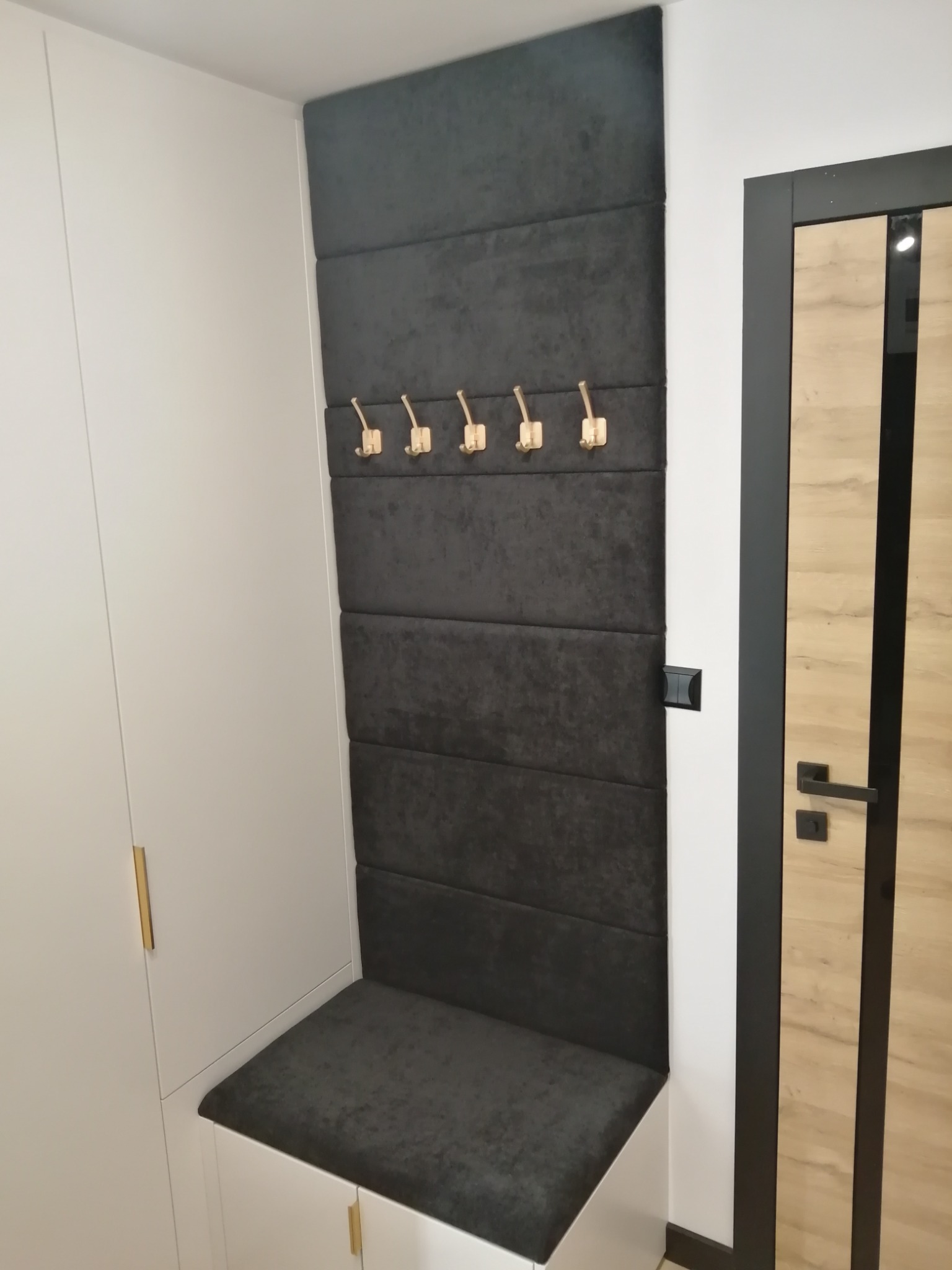 Tapicerowany panel ścienny z siedziskiem i złotymi haczykami w przedpokoju, obok biała szafa i drzwi z drewnianą okleiną.