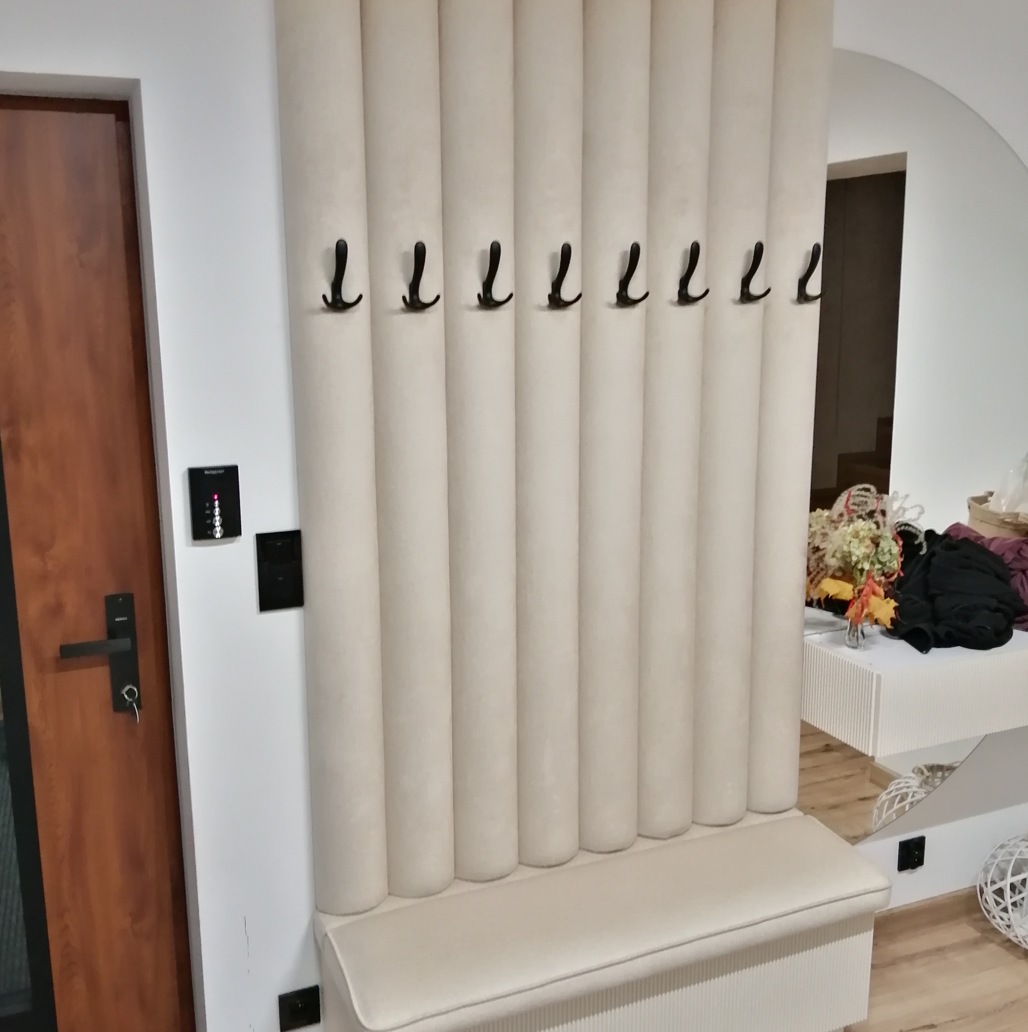 Tapicerowany panel ścienny z wieszakami i siedziskiem w przedpokoju, obok drzwi wejściowych z zamkiem elektronicznym.