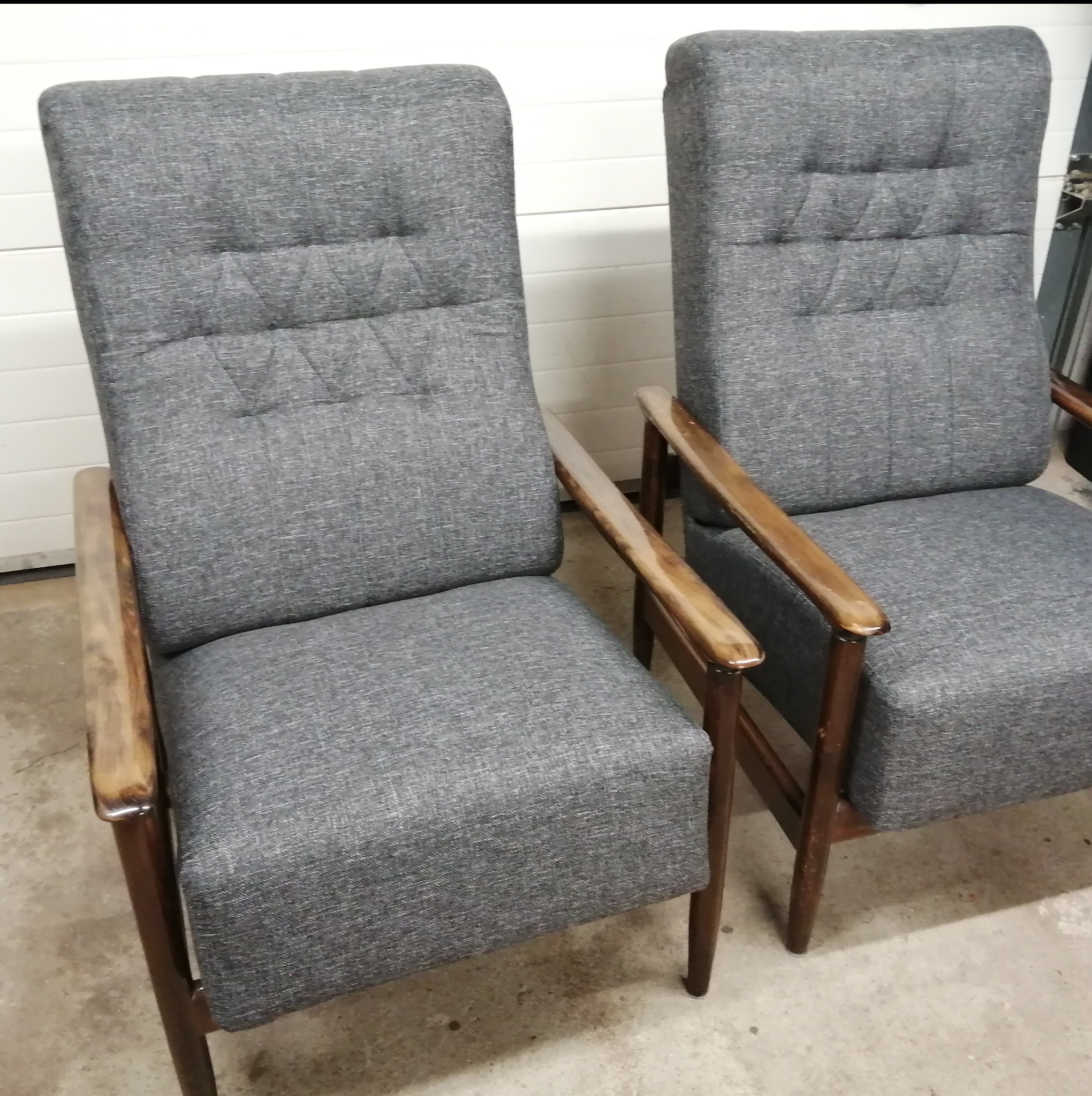 Dwa fotele tapicerowane szarą tkaniną z drewnianymi podłokietnikami, widoczne pionowe przeszycia na oparciach, ustawione na betonowej posadzce przed białą bramą garażową.