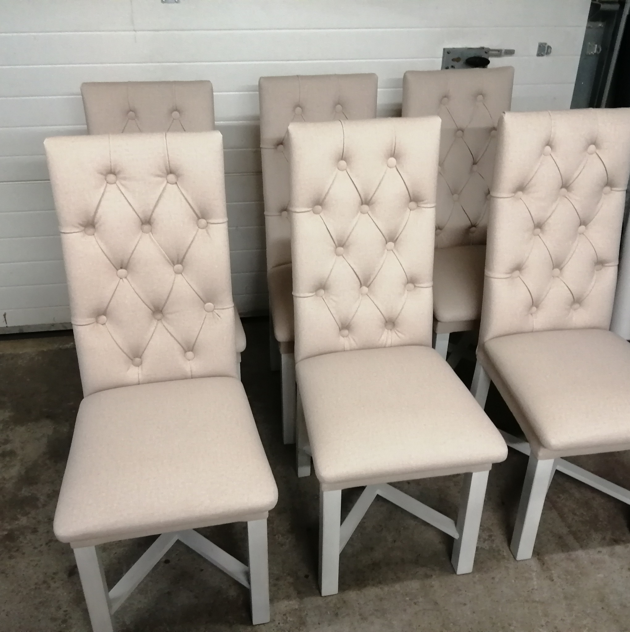 Rząd sześciu krzeseł z wysokim oparciem tapicerowanym w jasnym beżu, z pikowaniem w romby, stojących w pomieszczeniu przypominającym magazyn lub warsztat.