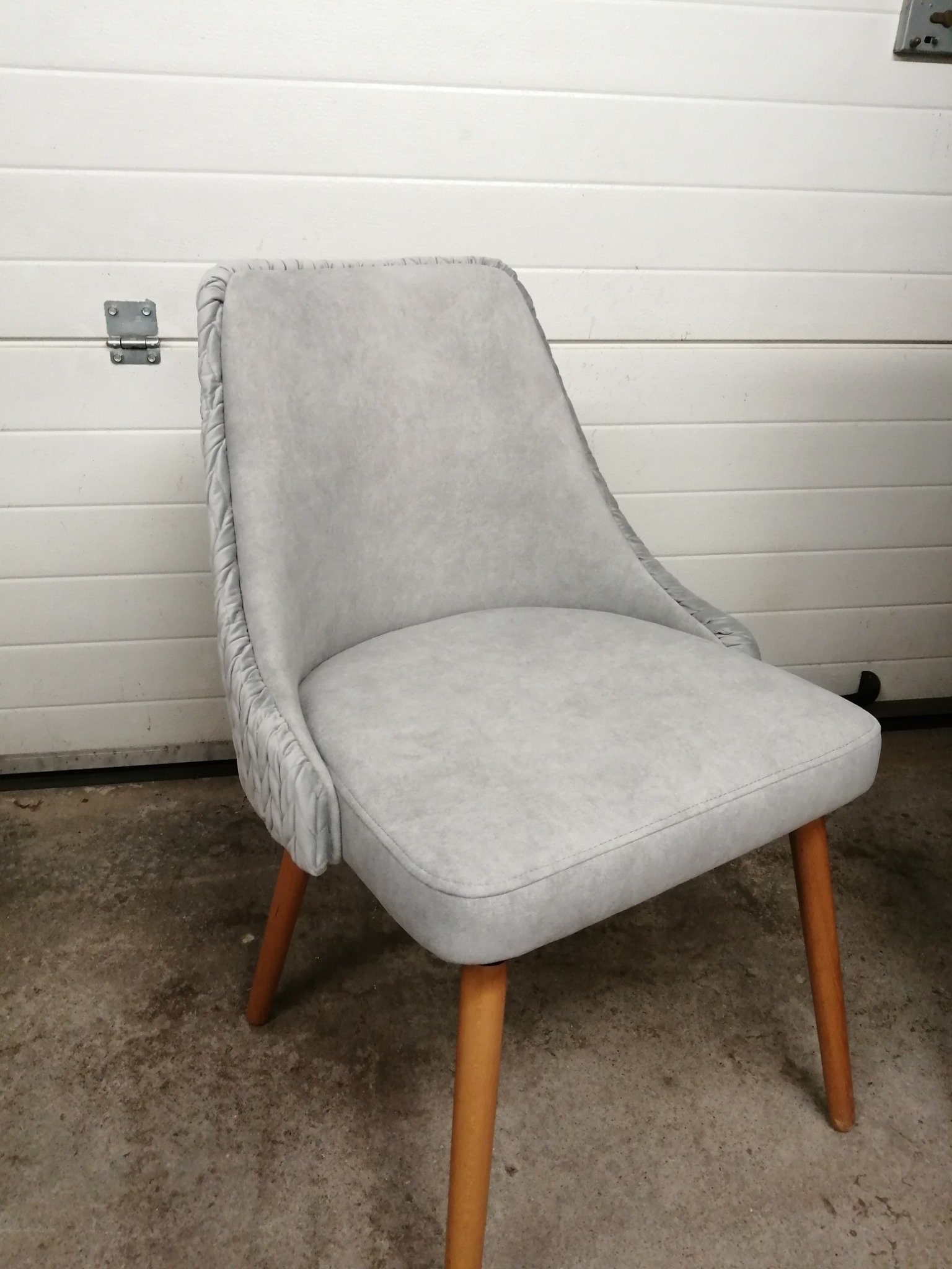 Szare krzesło tapicerowane z drewnianymi nogami na tle białej bramy garażowej, z charakterystycznym marszczeniem materiału na krawędzi oparcia.