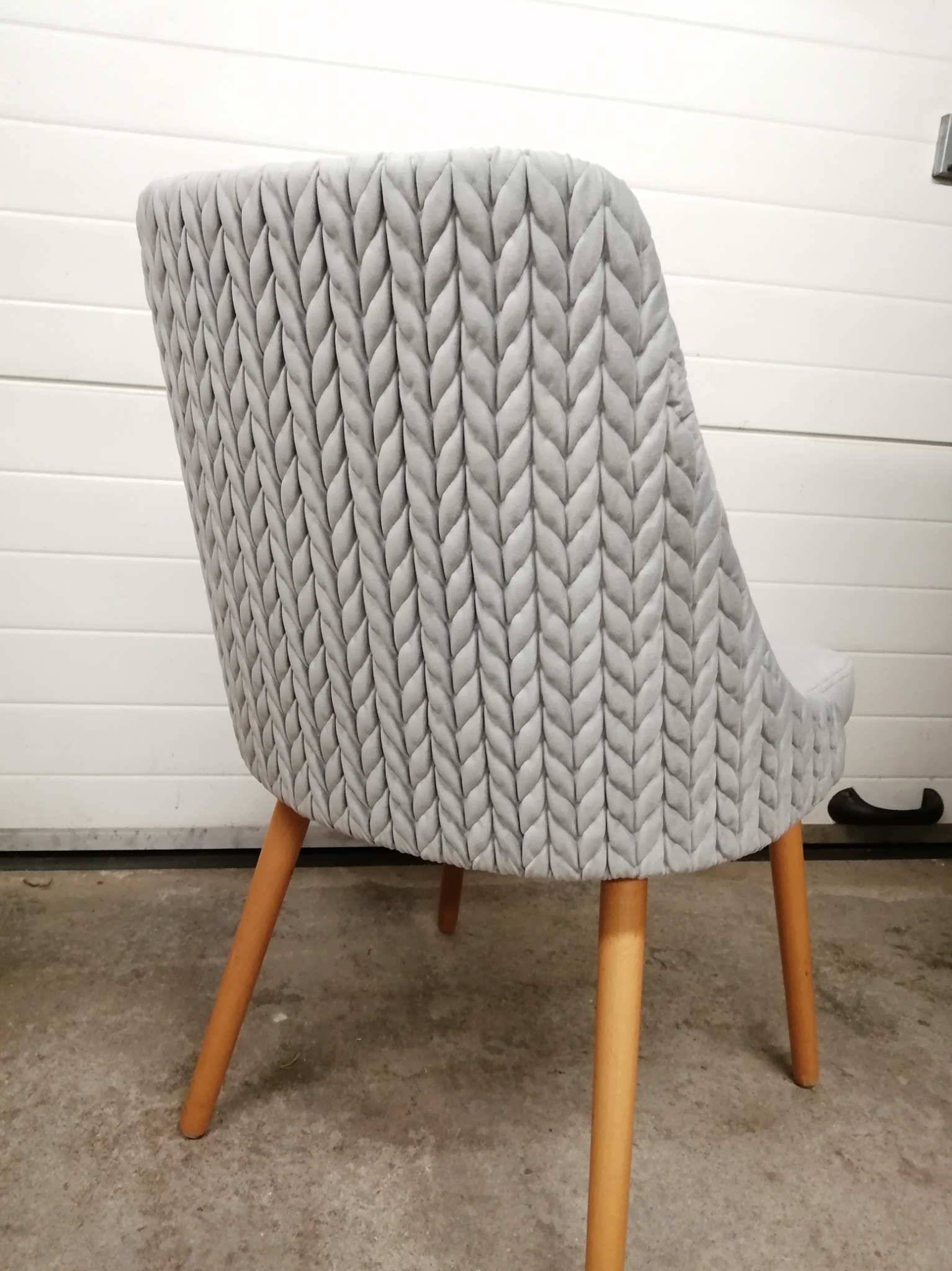 Szare krzesło tapicerowane z dekoracyjnym wzorem jodełki na oparciu, na drewnianych nogach, widok z tyłu na tle białej bramy garażowej.