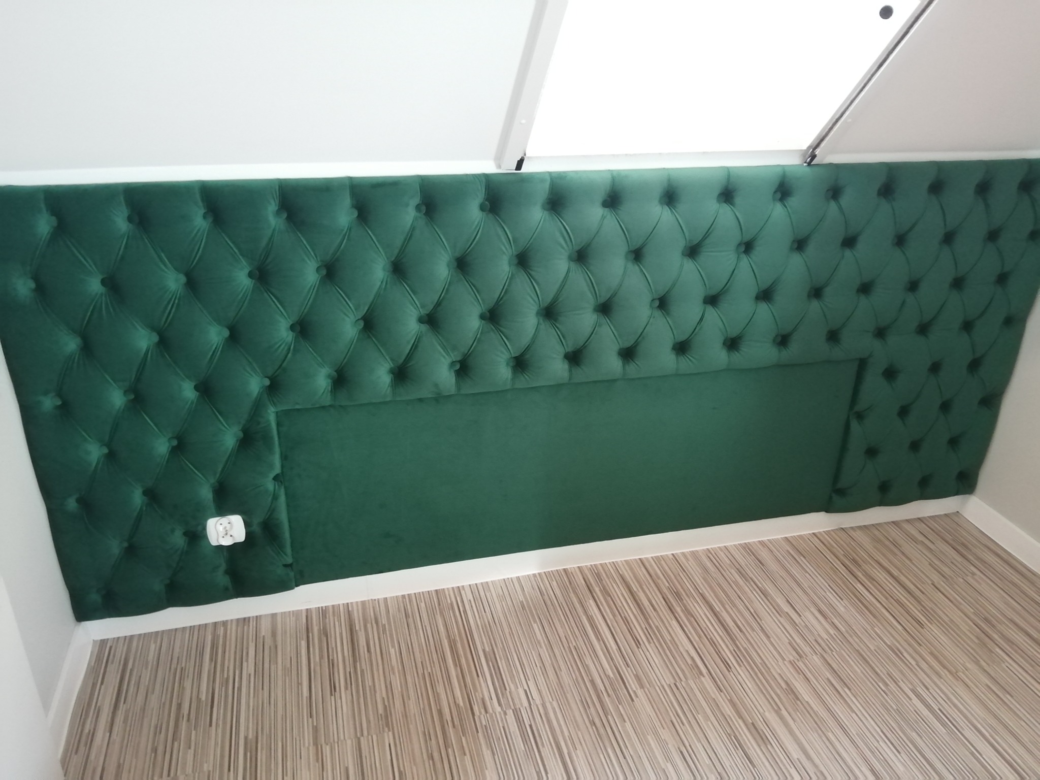 Szmaragdowozielony, pikowany panel tapicerowany na ścianie, częściowo pod skosem, z gładkim pasem na dole i widocznym gniazdkiem elektrycznym po lewej stronie.