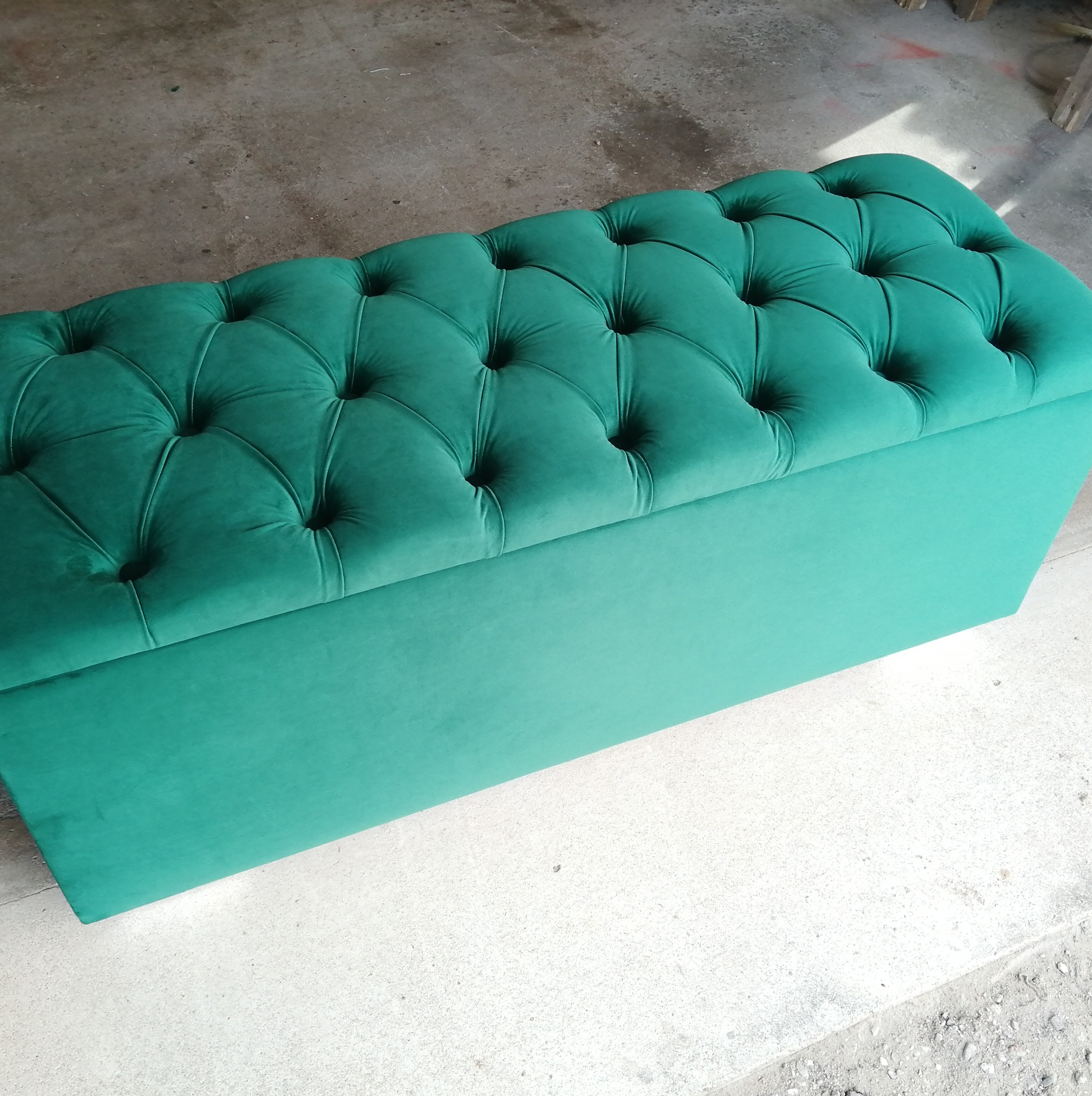 Turkusowa pufa tapicerowana z pikowaniem w romby, stojąca na szarym betonowym podłożu, widok z góry, delikatne oświetlenie.