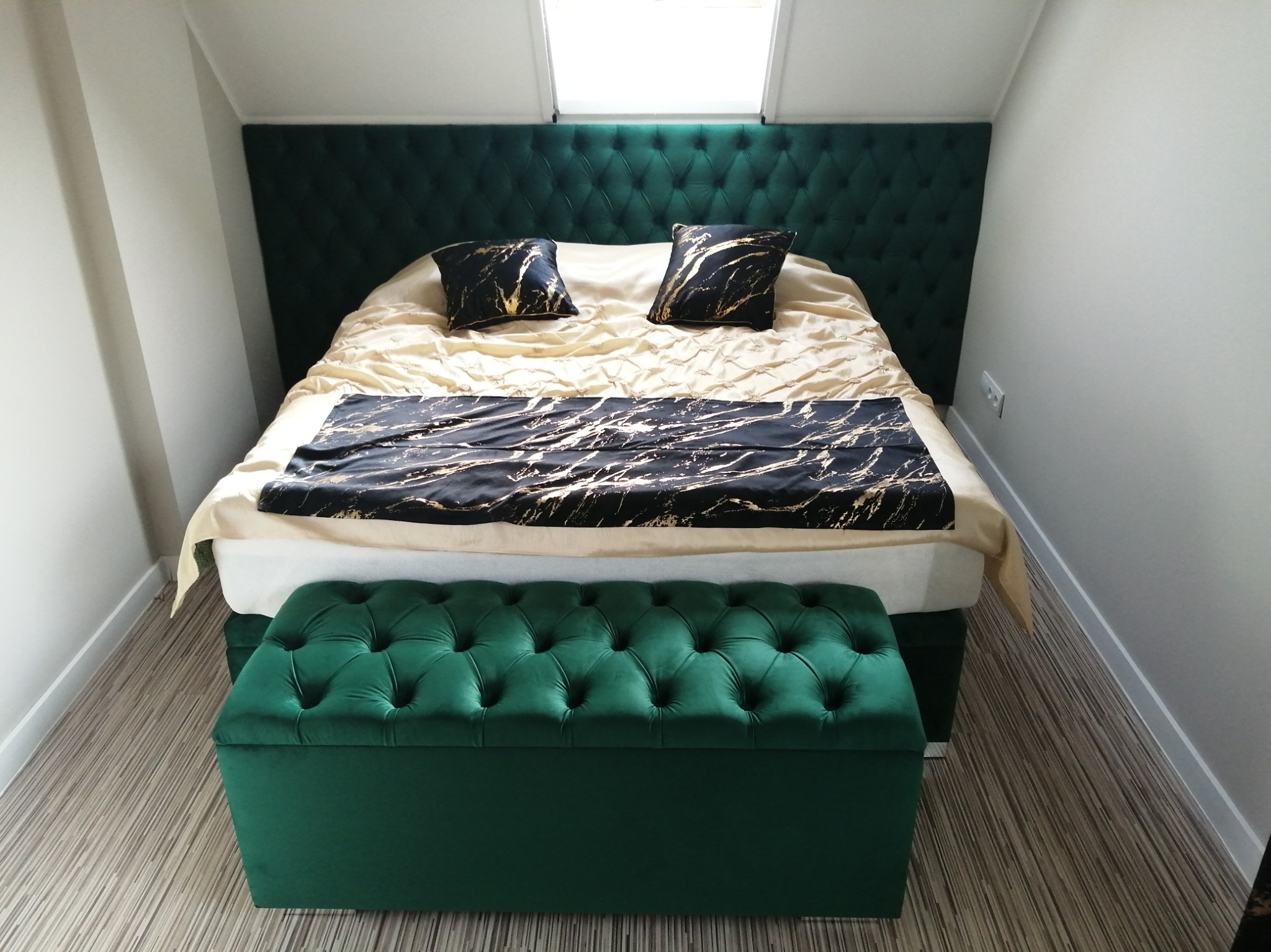 Szmaragdowy tapicerowany zagłówek łóżka z pikowaniem chesterfield, do kompletu pufa tapicerowana w tym samym stylu i kolorze, na łóżku beżowa pościel z czarno-złotym bieżnikiem i poduszkami.