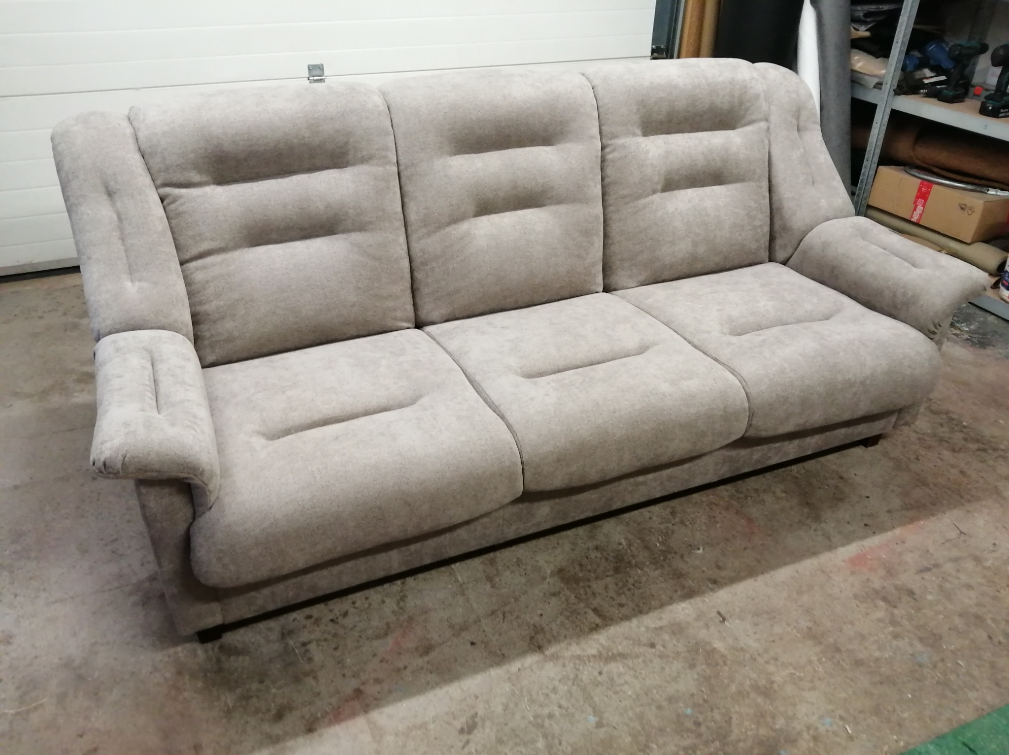 Trzyosobowa sofa w odcieniach beżu z charakterystycznymi przeszyciami na oparciach i siedziskach, sfotografowana w warsztatowym otoczeniu z widocznym regałem z narzędziami w tle.