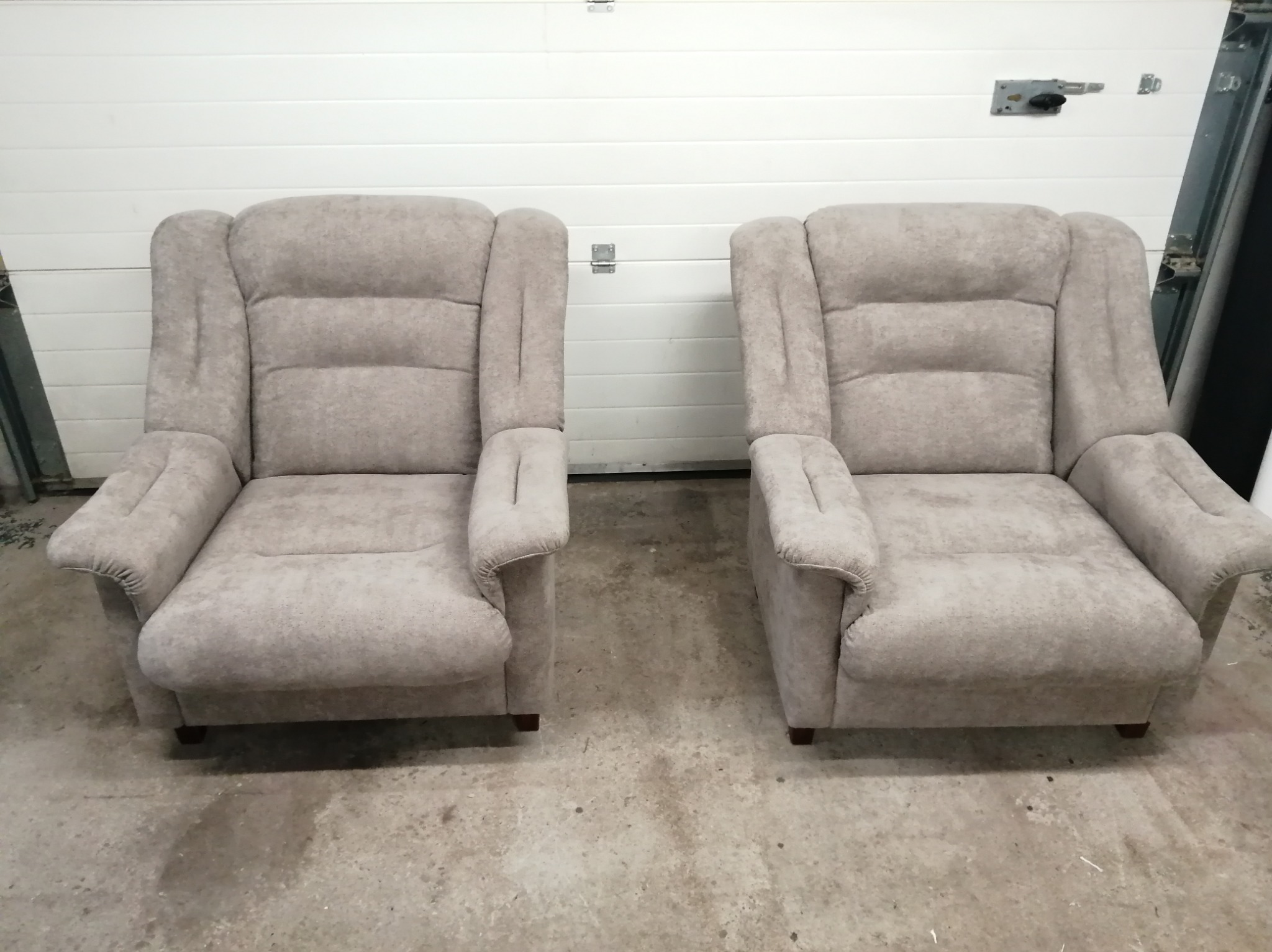 Dwa identyczne, beżowe fotele z podłokietnikami i trzema poziomymi przeszyciami na oparciu, ustawione obok siebie na betonowej podłodze, na tle białej bramy garażowej.