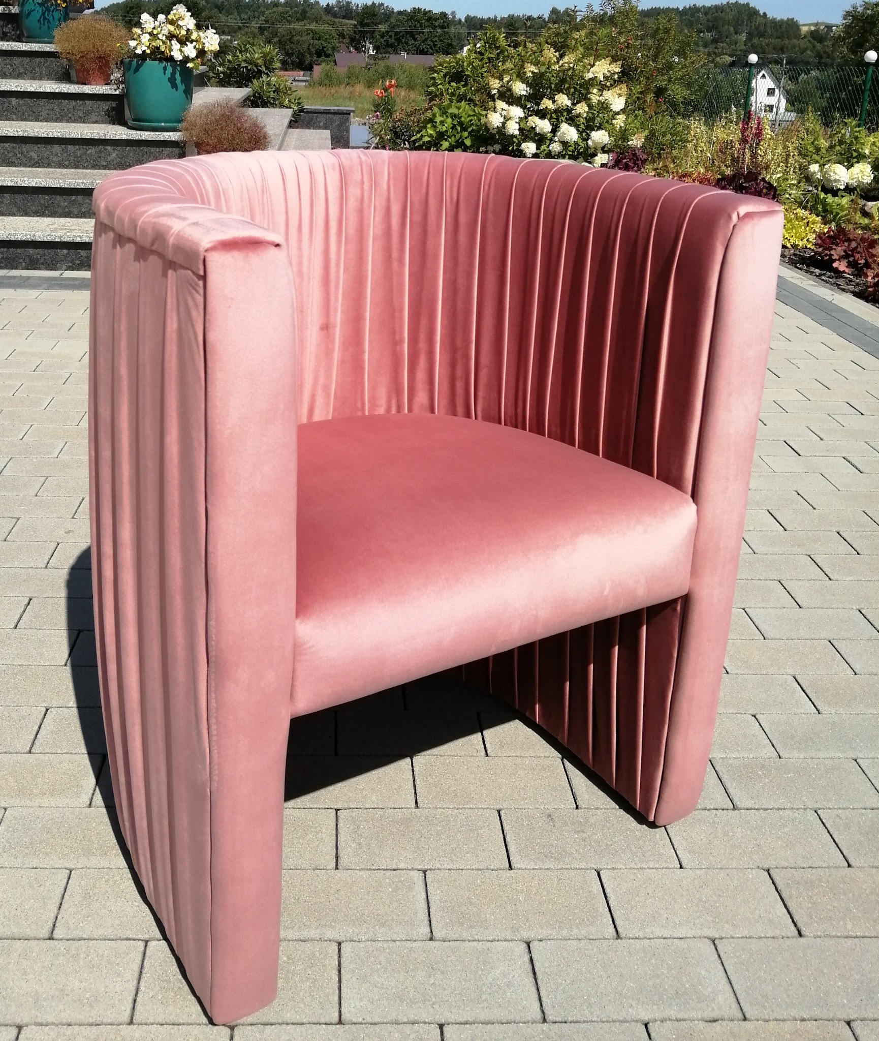 Różowe, welurowe krzesło z pionowymi przeszyciami, ustawione na brukowanej nawierzchni, z ogrodem i kwiatami w tle.