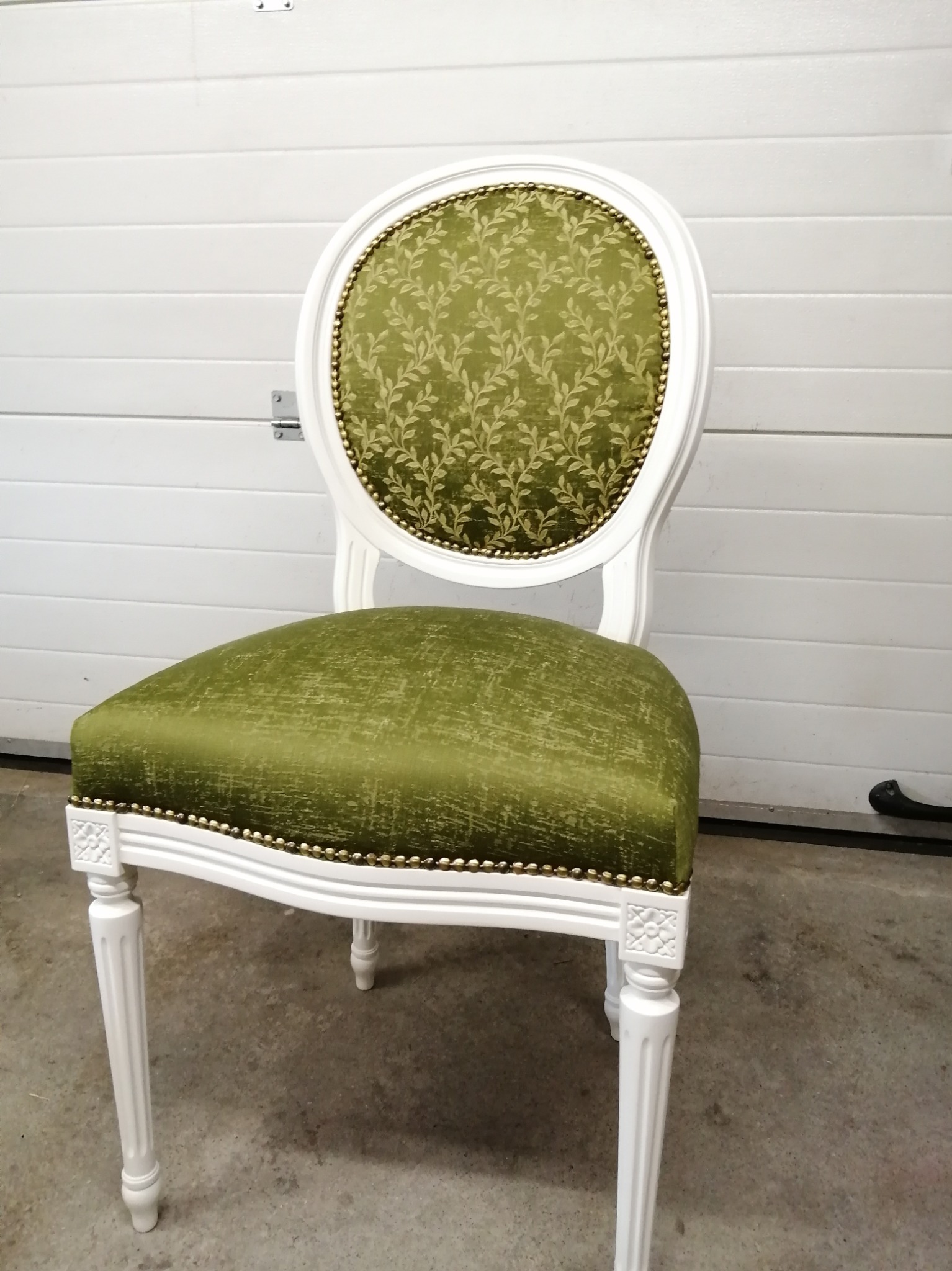 Krzesło w stylu Ludwika XVI z białą ramą i zieloną tapicerką z motywem roślinnym, wykończone ozdobnymi ćwiekami, na tle białej bramy garażowej.