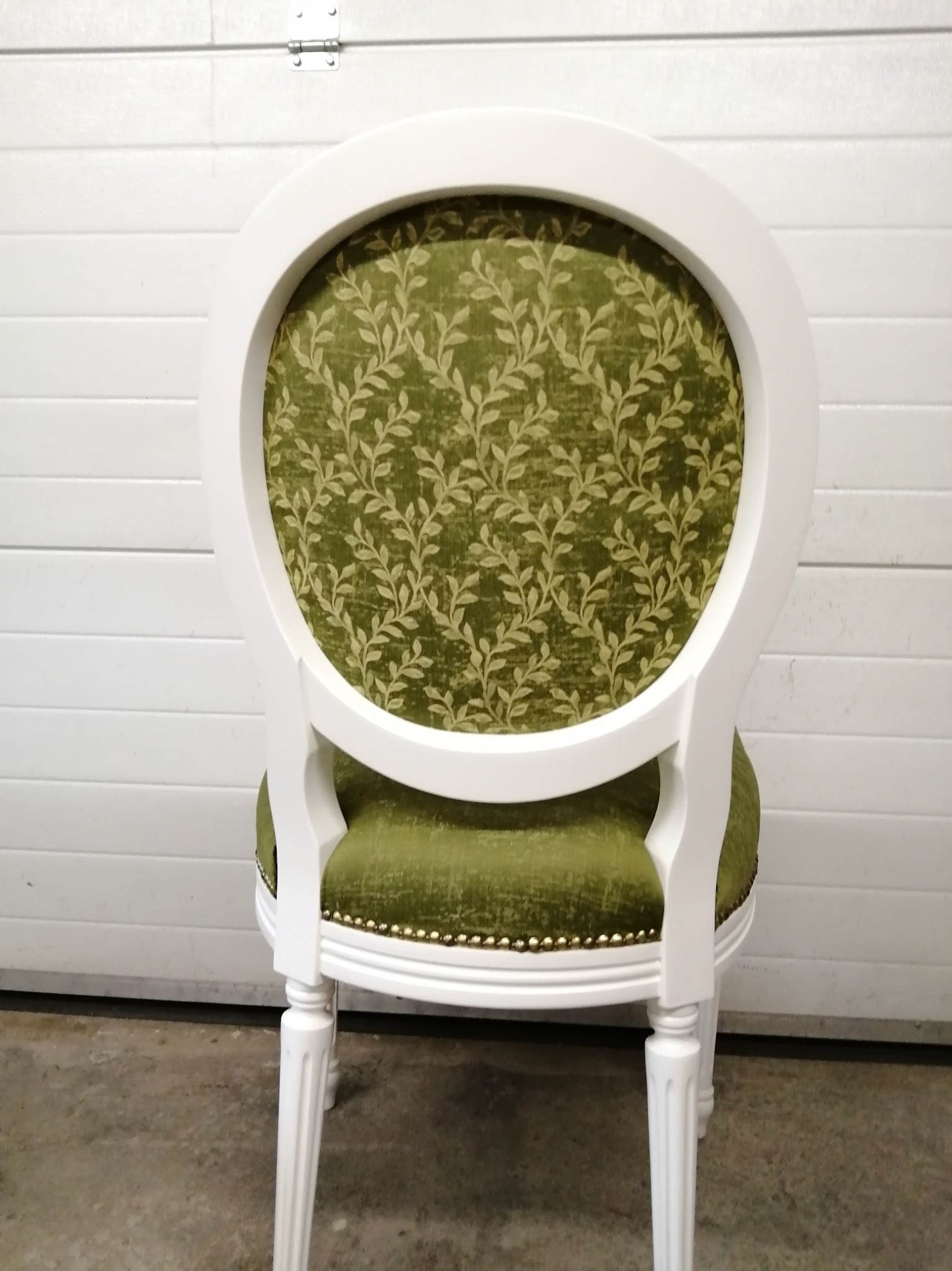 Krzesło z owalnym oparciem, białą ramą i zieloną tapicerką w roślinny wzór, widoczne od tyłu na tle białej, garażowej bramy.