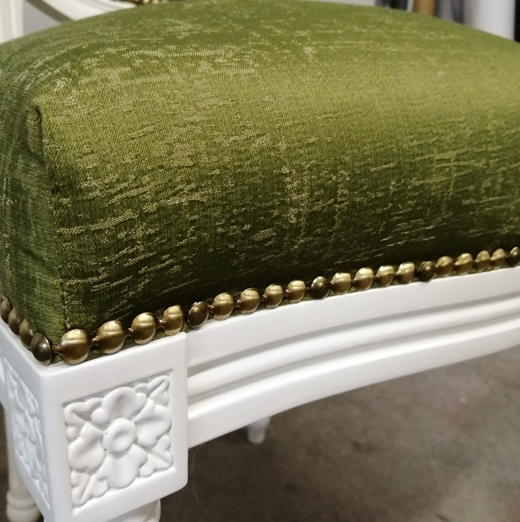 Fragment odnowionego tapicerowanego siedziska z zielonego materiału, wykończonego ozdobnymi, mosiężnymi gwoździami na białej, rzeźbionej ramie.