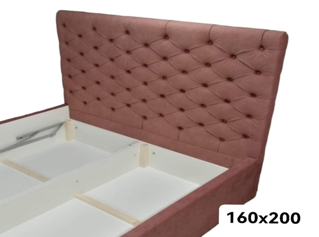 Różowe łóżko tapicerowane z zagłówkiem w romby, widoczny mechanizm podnoszenia stelaża i pusta przestrzeń na pościel, wymiary 160x200 cm.