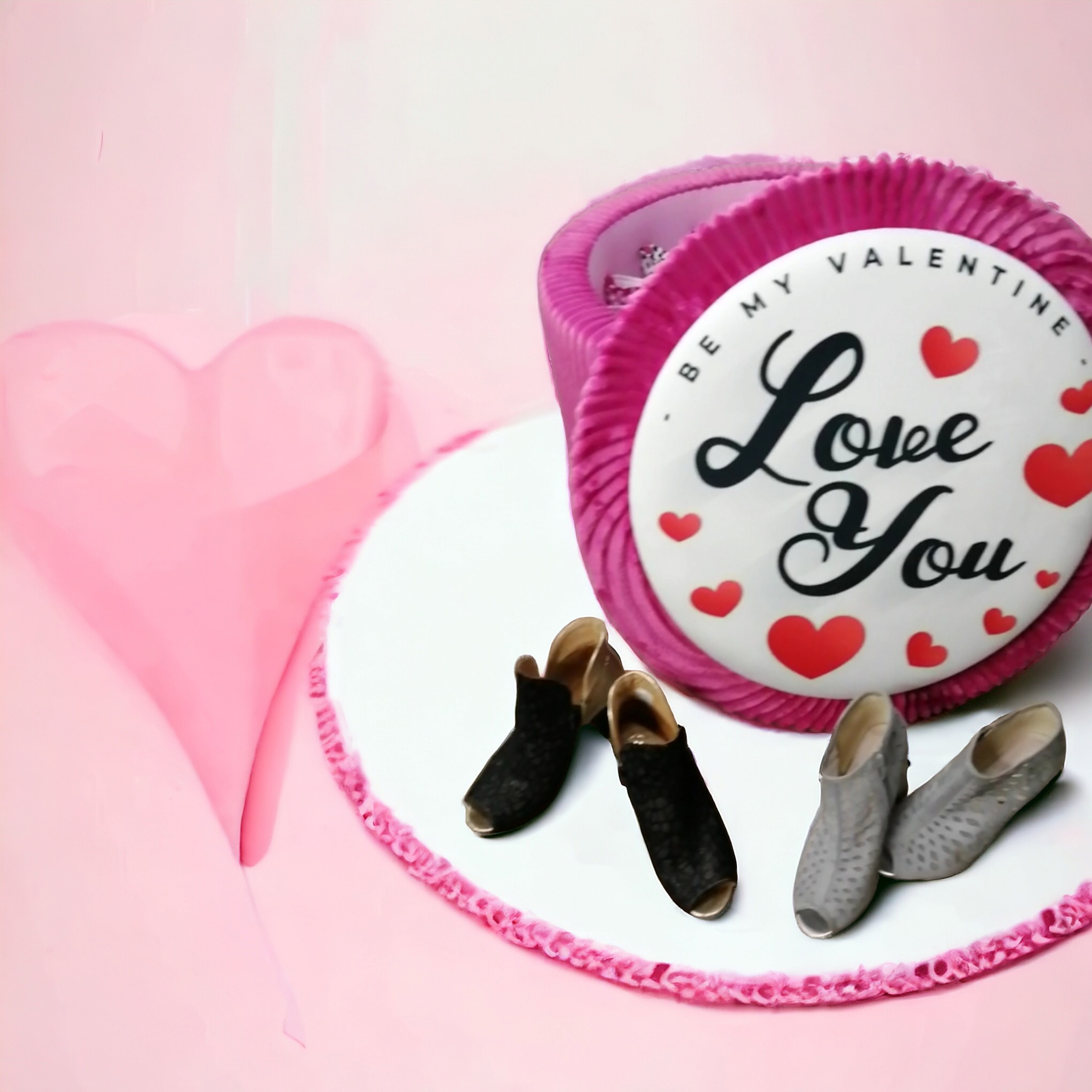 Różowe pudełko w kształcie pierścienia z napisem 'Love You' i serduszkami, otoczone dwoma parami damskich butów z odkrytymi palcami na białym podeście z różowym brzegiem, obok papierowe serce.