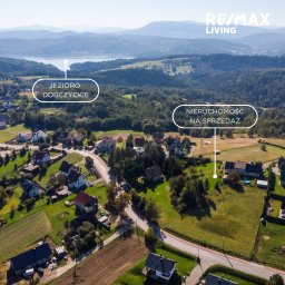 Ewelina Sendecka SeeWell HOME - Panoramiczny widok z lotu ptaka na jezioro Dobczyckie i okoliczne domy, oznaczona nieruchomość z czerwonym samochodem na sprzedaż, logo RE/MAX Living w prawym górnym rogu.