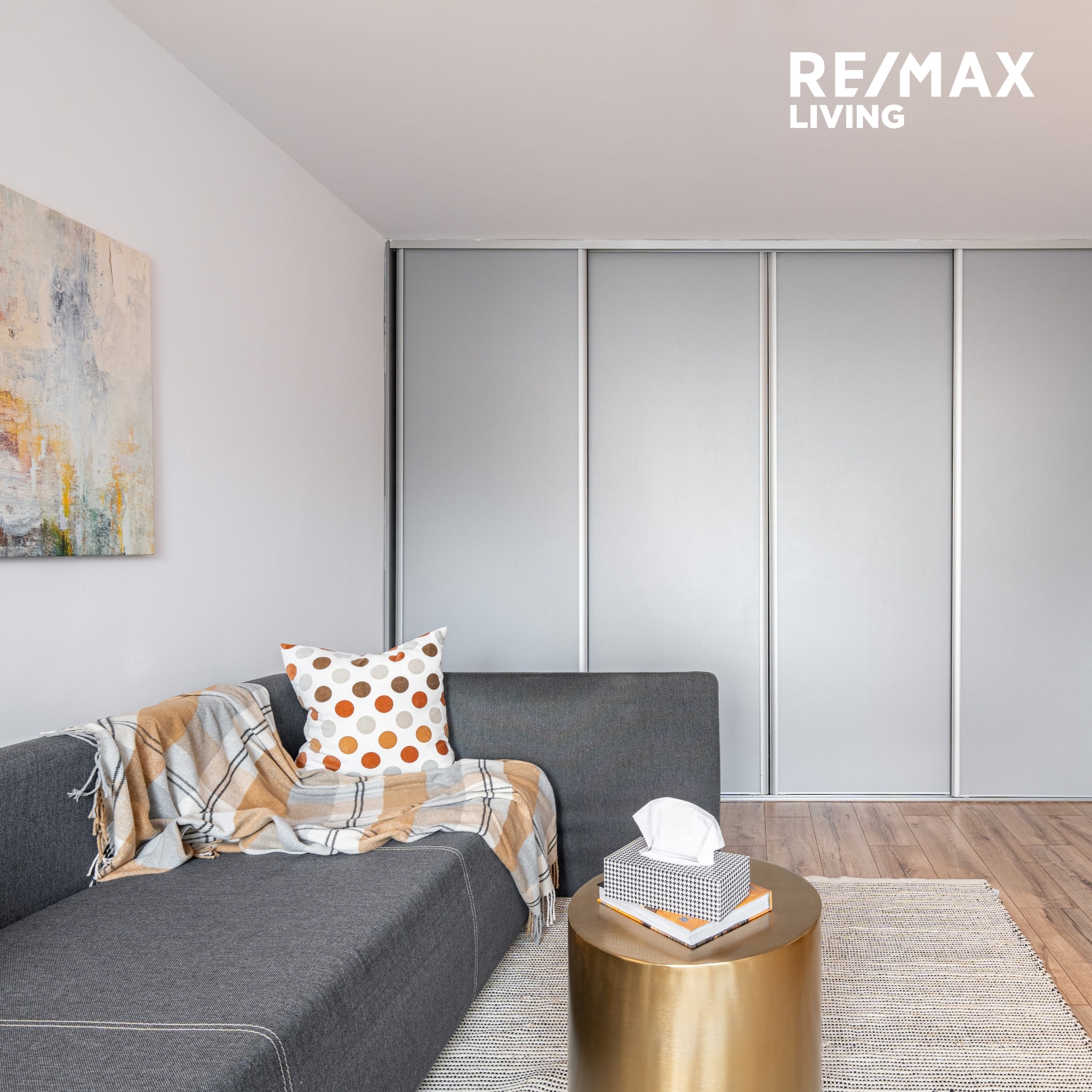 Nowoczesny salon z szarą sofą, pledem w kratę, poduszką w kropki i złotym stolikiem na tle przesuwnych drzwi szafy i abstrakcyjnego obrazu.