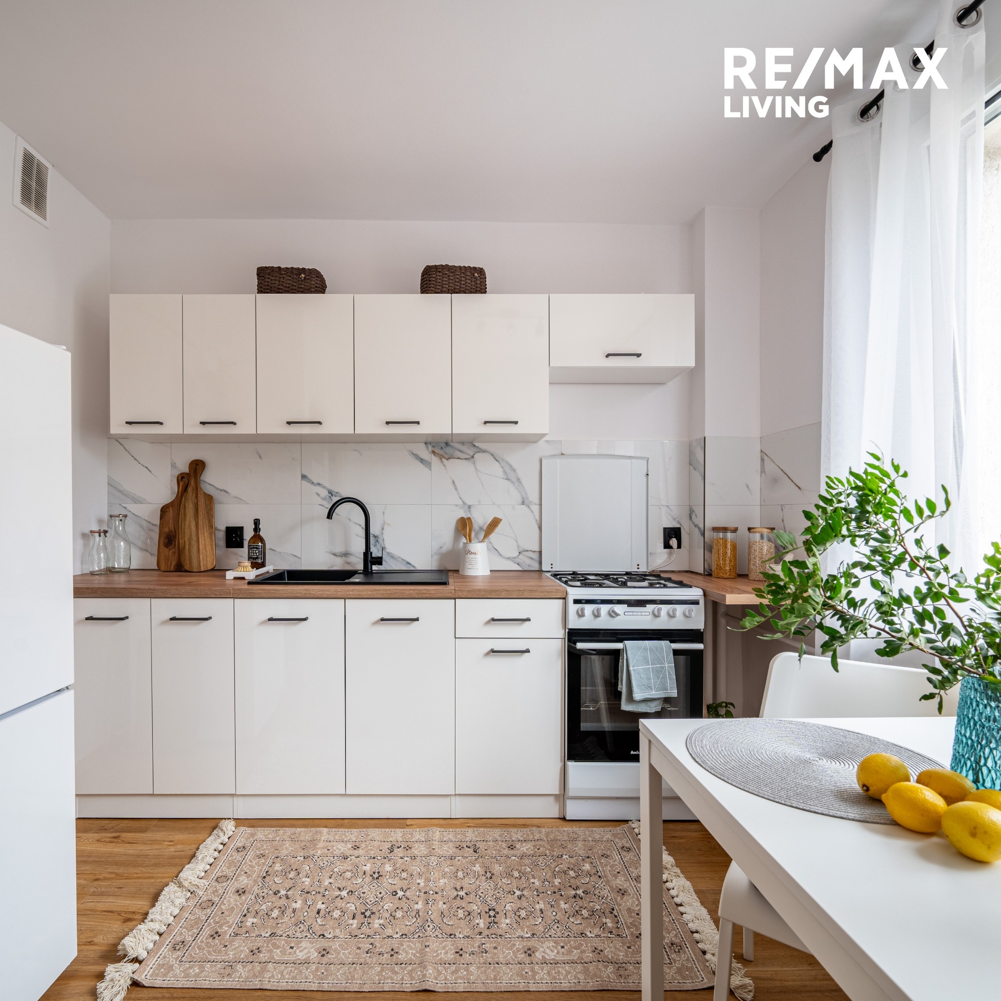 Jasna kuchnia z białymi szafkami, drewnianym blatem i marmurowymi płytkami na ścianie, dywan w stylu vintage na podłodze i stół z cytrynami, oświetlona naturalnym światłem, logo RE/MAX Living...