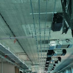 HYDROTECH Bogdan Wrona, Paweł Lis spółka cywilna - Instalacja rur i przewodów wentylacyjnych pod betonowym sufitem w budynku w trakcie budowy, widoczne elementy mocujące i fragmenty systemów wentylacyjnych.