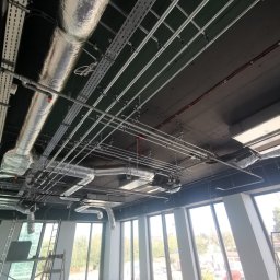 HYDROTECH Bogdan Wrona, Paweł Lis spółka cywilna - Widok z dołu na sufit w nowym budynku z widocznymi instalacjami: rurami wentylacyjnymi w srebrnej izolacji, metalowymi korytkami kablowymi i rurami instalacji przeciwpożarowej, z oknami na zewnątrz.