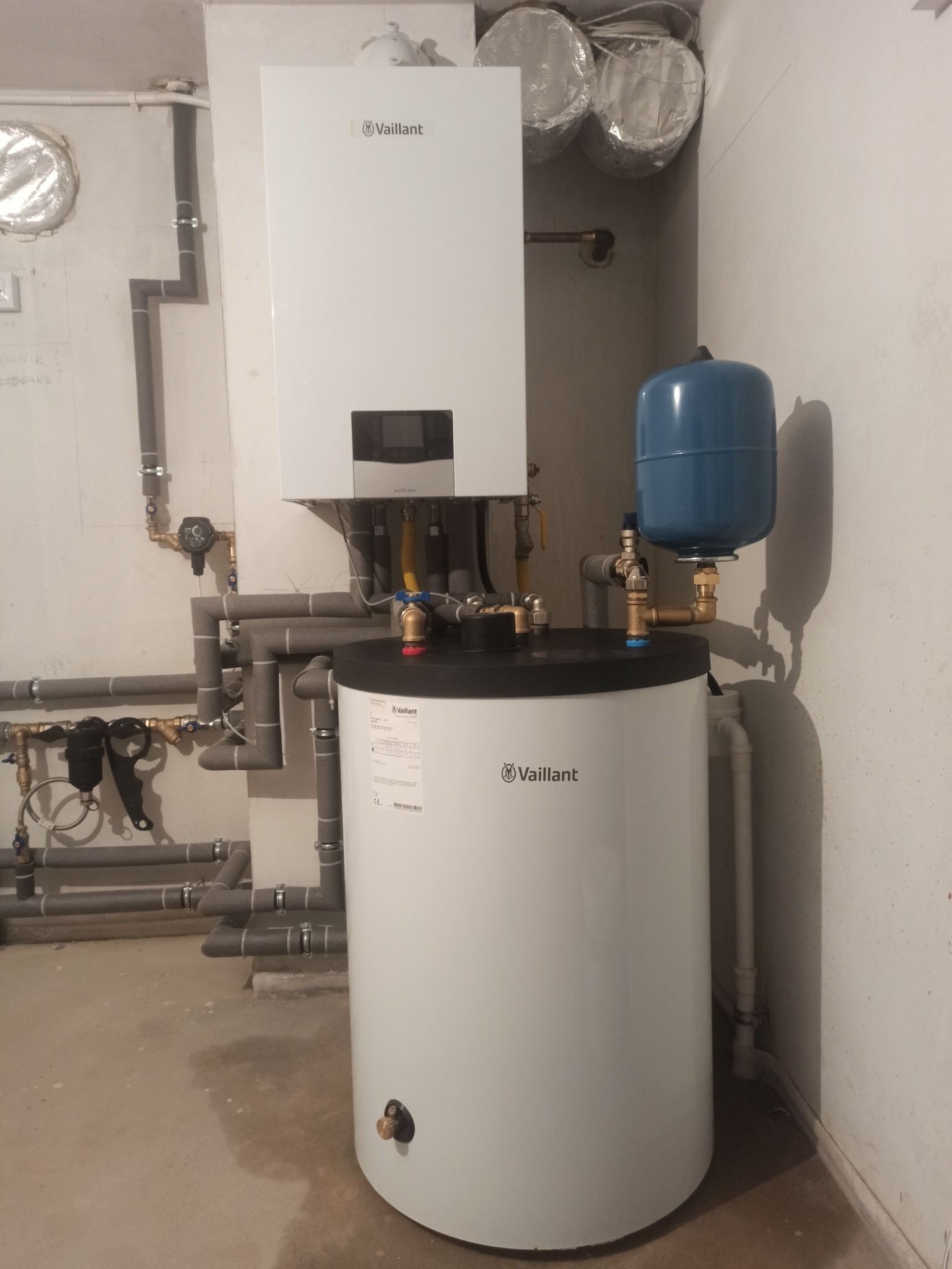 Biały piec gazowy Vaillant z podłączonym zasobnikiem wody i niebieskim naczyniem wzbiorczym, widoczne szare rury instalacji grzewczej z izolacją.