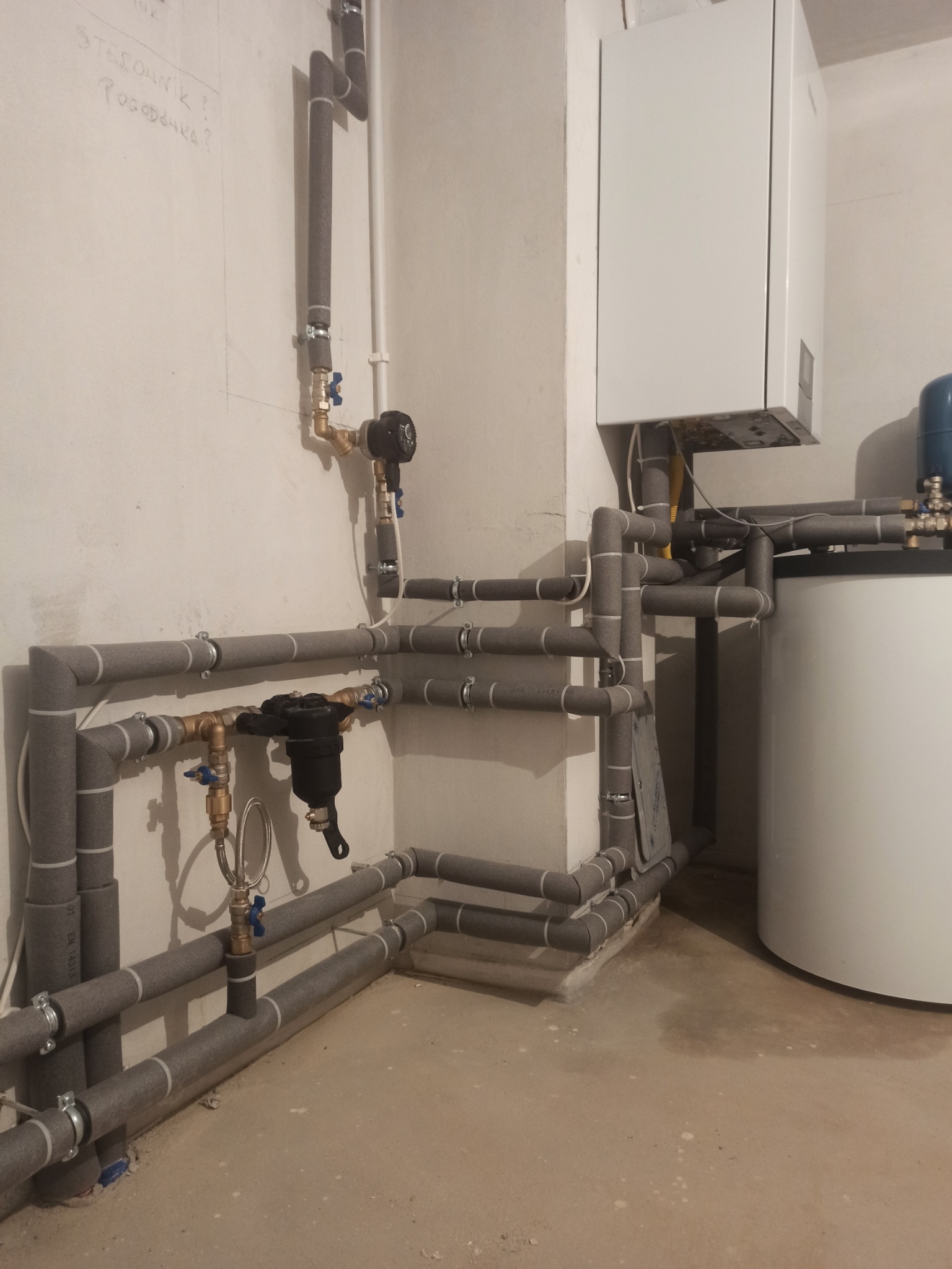 Kotłownia z nową instalacją centralnego ogrzewania, widoczne rury w izolacji, zawory, filtr oraz piec gazowy wiszący na ścianie, na ścianie notatki o sterowniku.