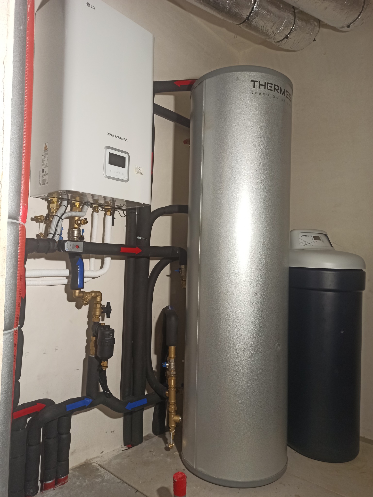 Biały piec grzewczy LG Therma V wiszący na ścianie, obok srebrny zbiornik Thermes Green Solution i czarny filtr do wody, rury z izolacją w kolorze czarnym z oznaczeniami kierunku przepływu wody...