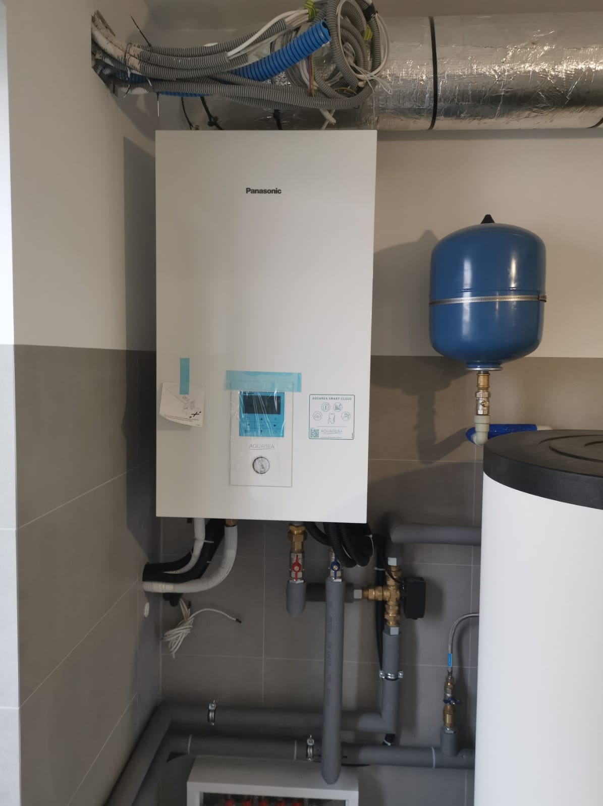 Zainstalowany piec gazowy Panasonic Aquarea z niebieskim zbiornikiem wyrównawczym, widoczne rury instalacji grzewczej i przyłącza wodne na tle szarych płytek.
