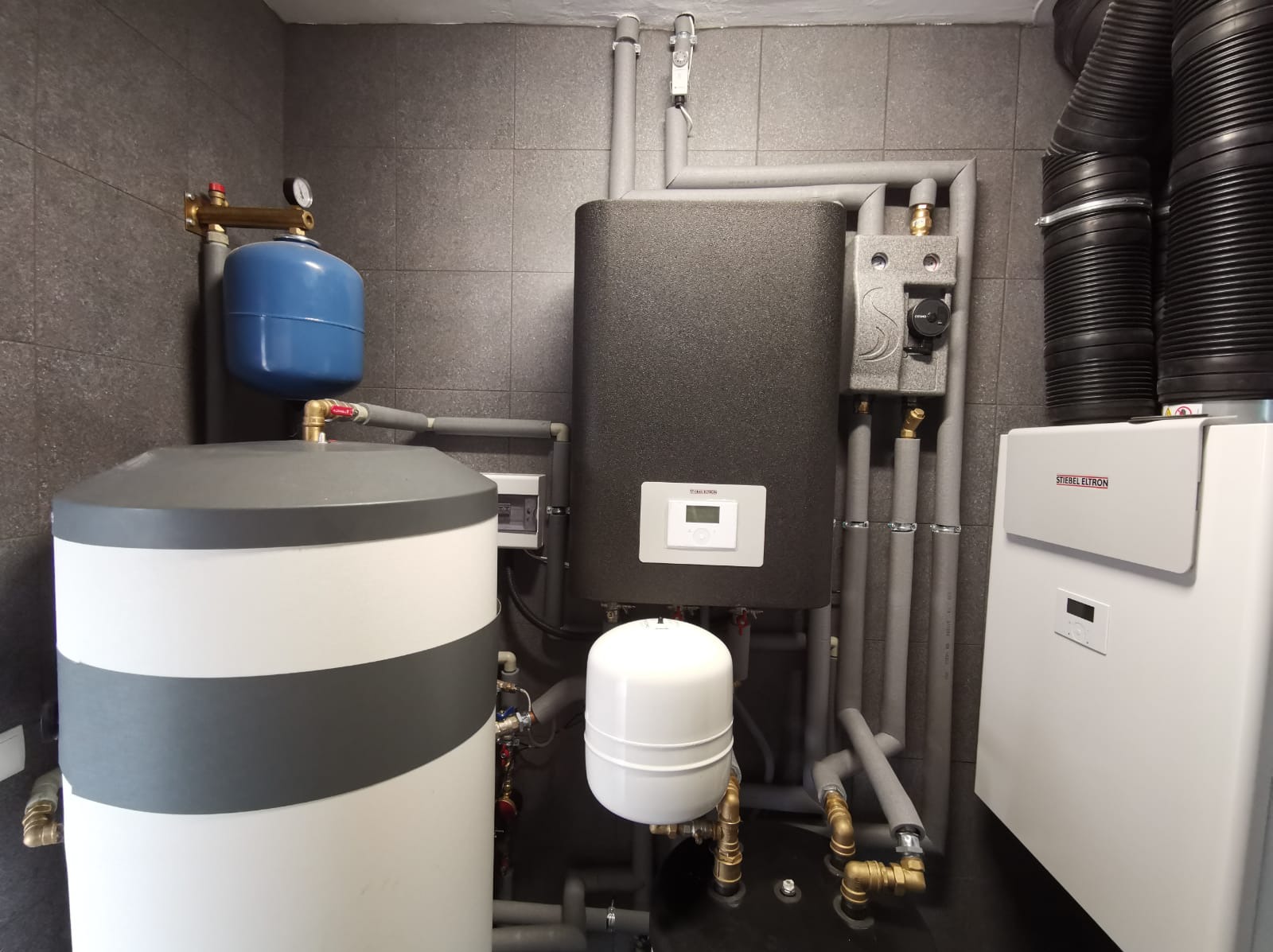 Kotłownia z piecem Stielbel Eltron, zbiornikiem akumulacyjnym z szarym pasem, orurowaniem w izolacji oraz szarą płytką na ścianach.