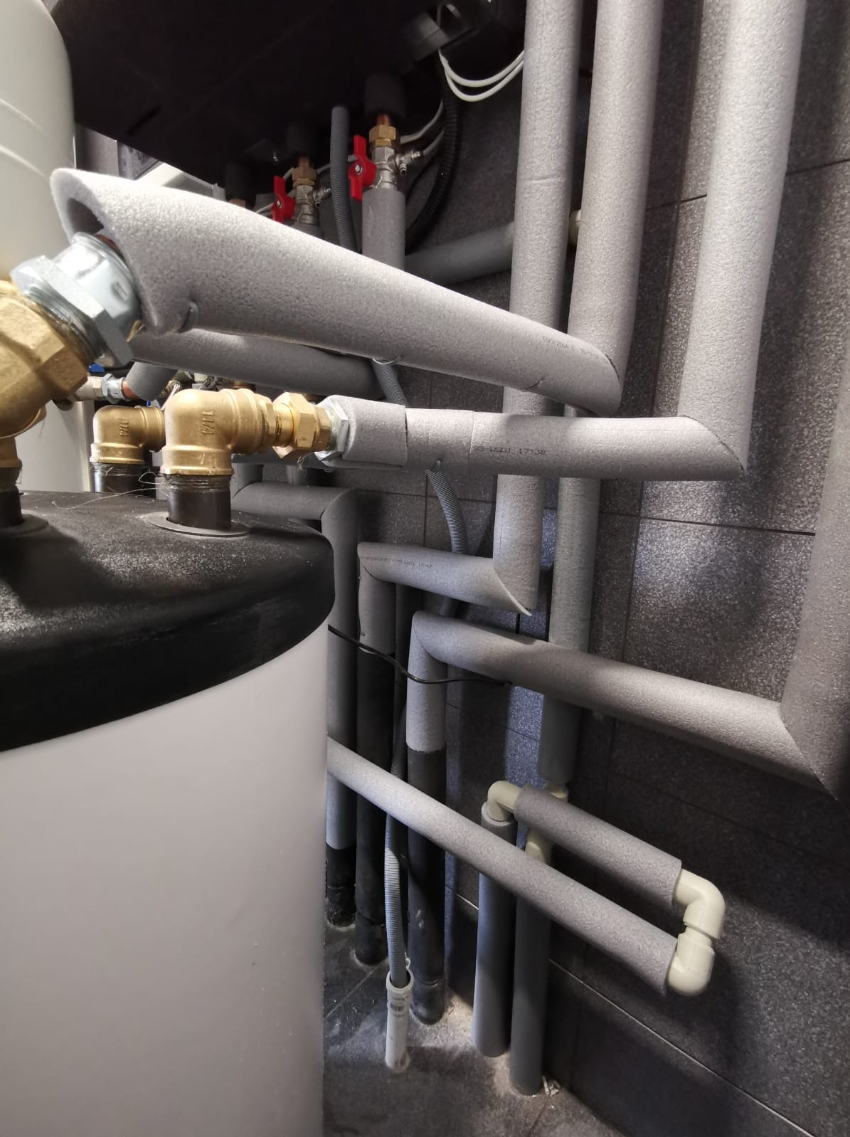 Szare, zaizolowane rury instalacji grzewczej z licznymi złączkami, zaworami i elementami montażowymi na tle ciemnej, szarej ściany wyłożonej płytkami. Widoczny fragment białego zasobnika.