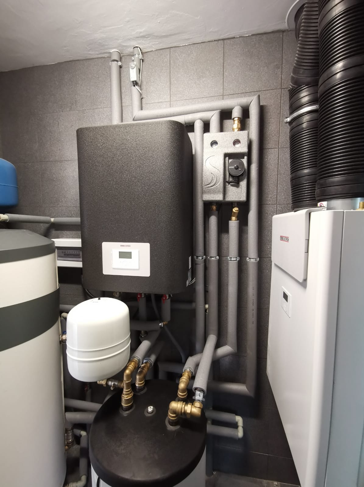 Zestaw urządzeń grzewczych w kotłowni: zbiornik buforowy, pompa ciepła i inne elementy instalacji, rury zaizolowane szarą pianką.