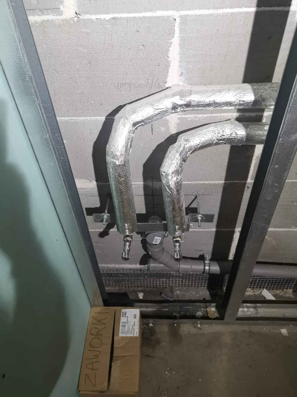 Rury grzewcze owinięte folią izolacyjną, zamontowane na ścianie z surowych bloczków, widoczne elementy mocujące i fragment podłogi z kratką wentylacyjną, karton z napisem 'ZAWORKI' w lewym dolnym...