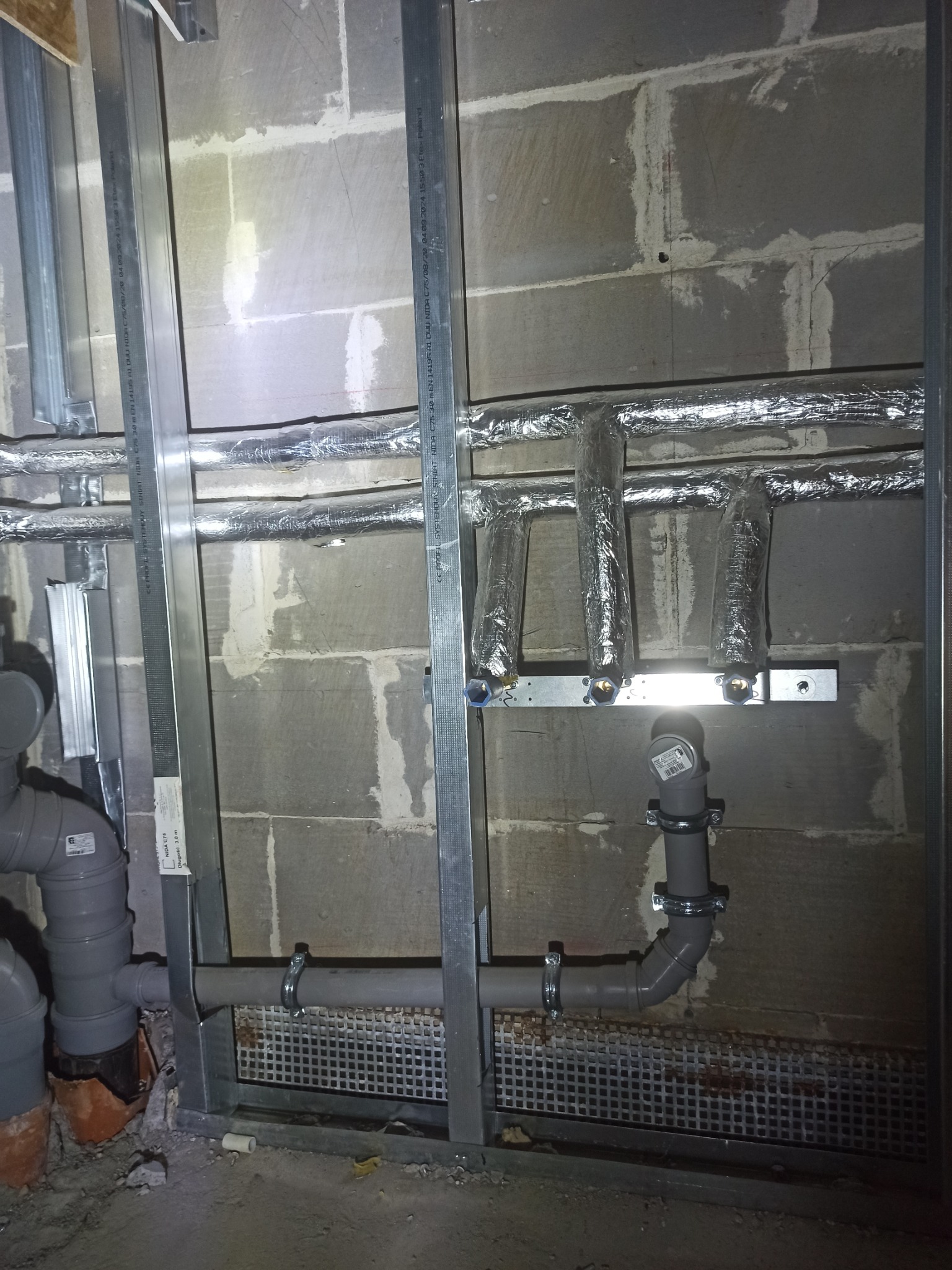 Instalacja rur kanalizacyjnych i grzewczych w surowym stanie, rury izolowane folią aluminiową, widoczne metalowe profile konstrukcyjne, ściana z pustaków betonowych w tle.