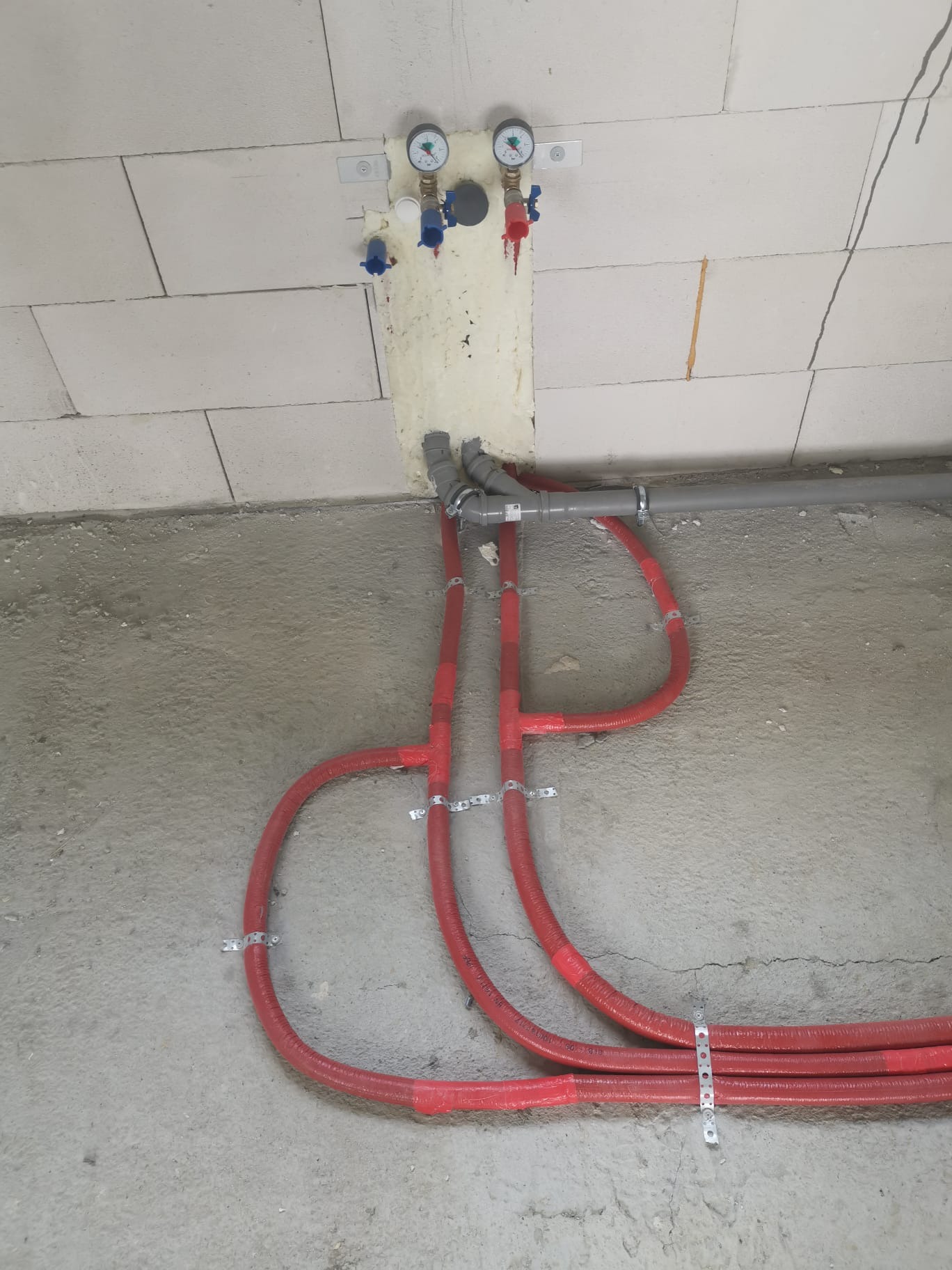 Czerwone rury grzewcze, przymocowane metalowymi uchwytami do surowego betonowego podłoża, z szarymi rurami odpływowymi i manometrami na ścianie z białych bloczków.
