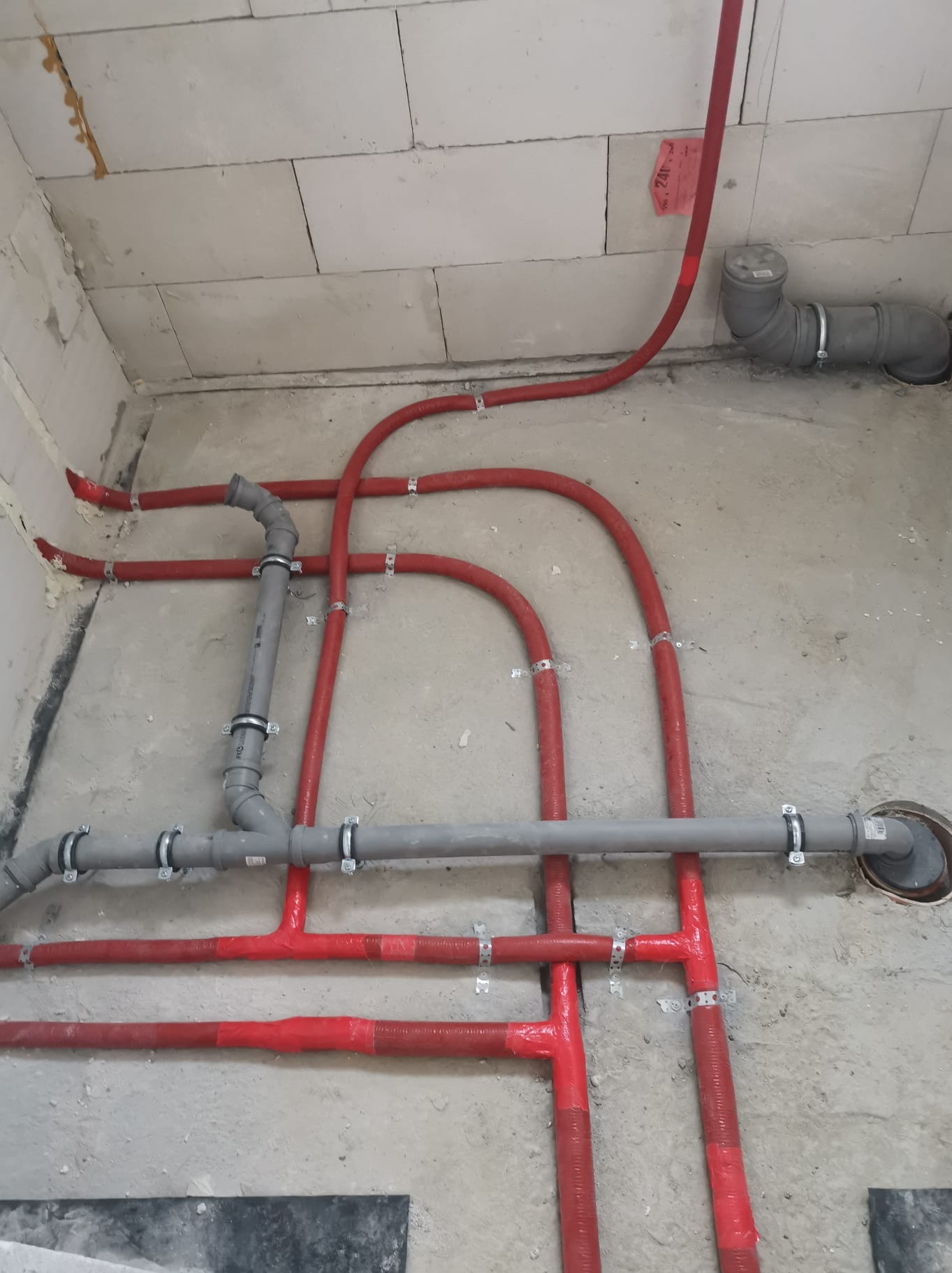 Czerwone rury ogrzewania podłogowego i szare rury kanalizacyjne zamontowane na surowym betonie w Skawinie, widok z góry.