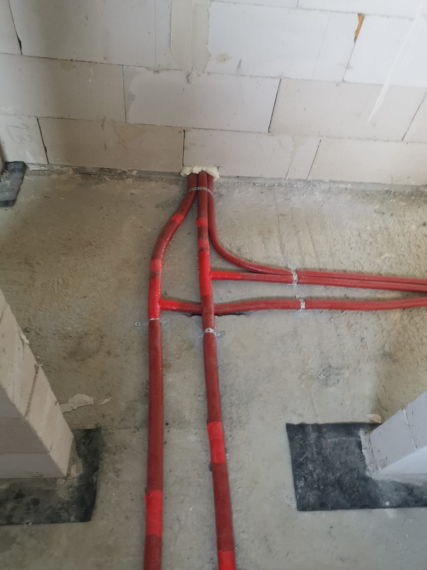 Czerwone rury instalacyjne, zabezpieczone pianką montażową przy ścianie z betonu komórkowego, przymocowane do podłoża metalowymi uchwytami.