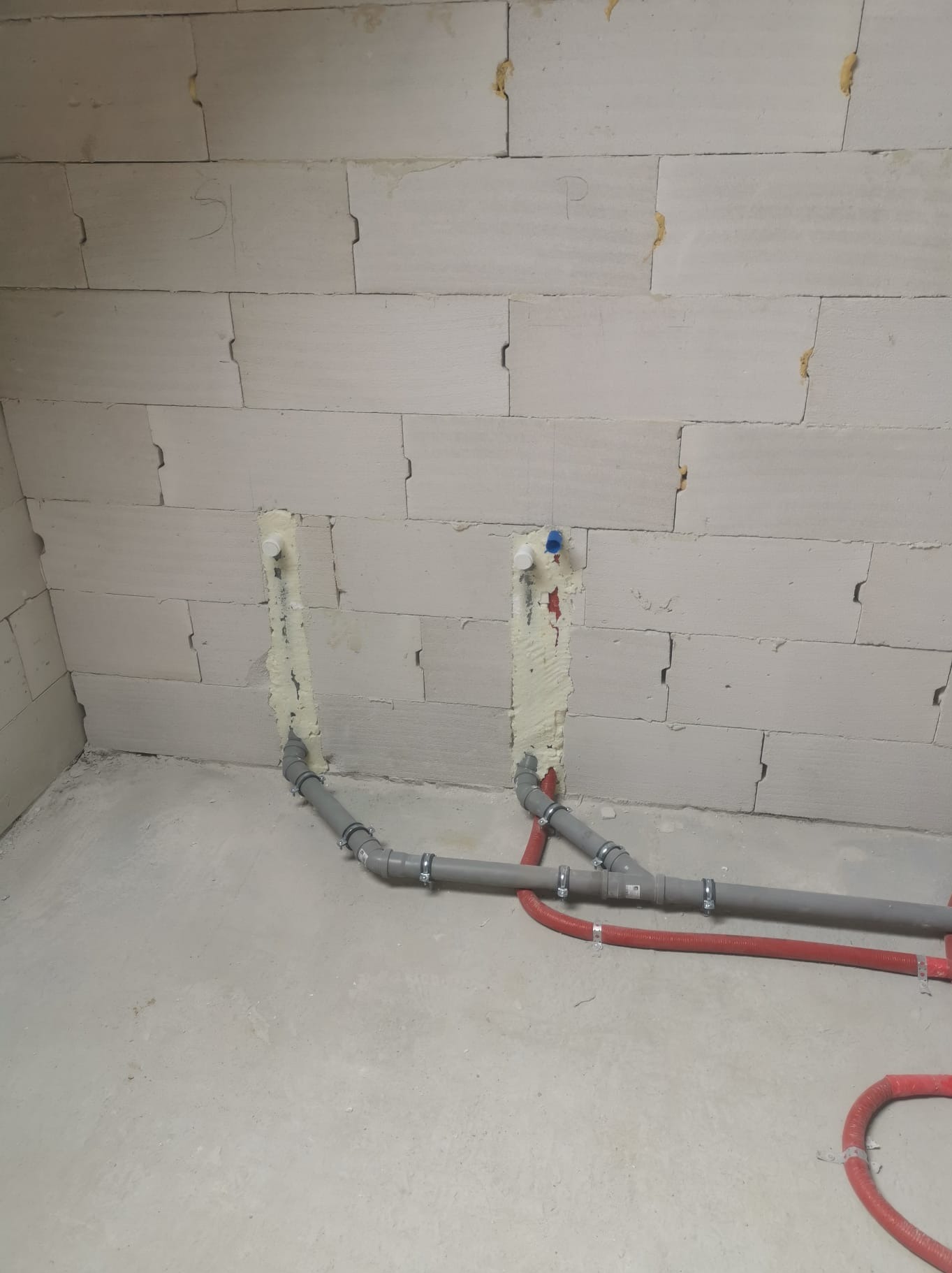 Surowe wnętrze w trakcie prac: szare i czerwone rury instalacji wodnej i kanalizacyjnej zamocowane do ściany z białych bloczków, zabezpieczone pianką montażową.