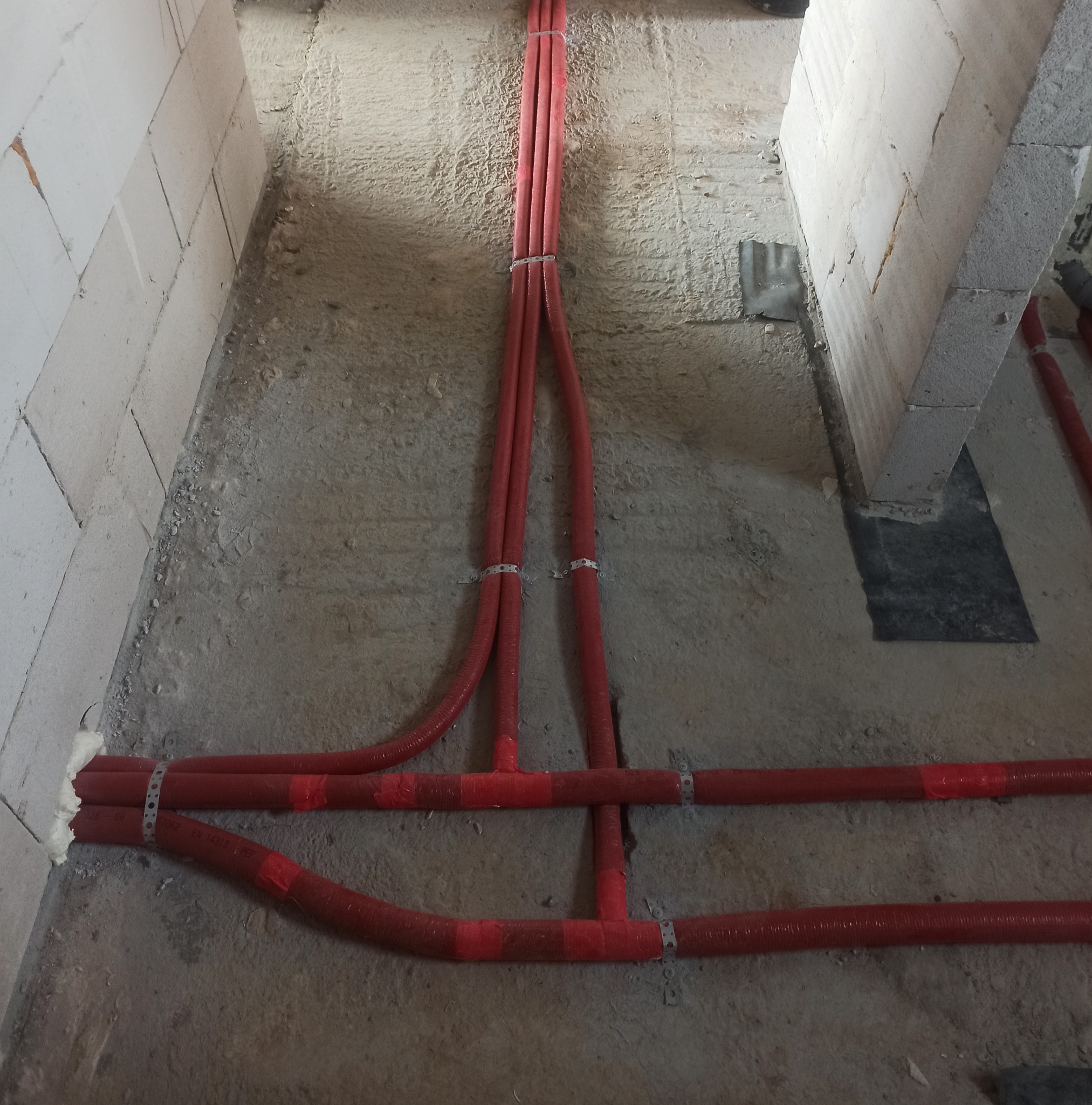 Czerwone, karbowane rury instalacyjne ułożone na surowym betonowym podłożu, przymocowane metalowymi obejmami, widoczne fragmenty ścian z białego betonu komórkowego.