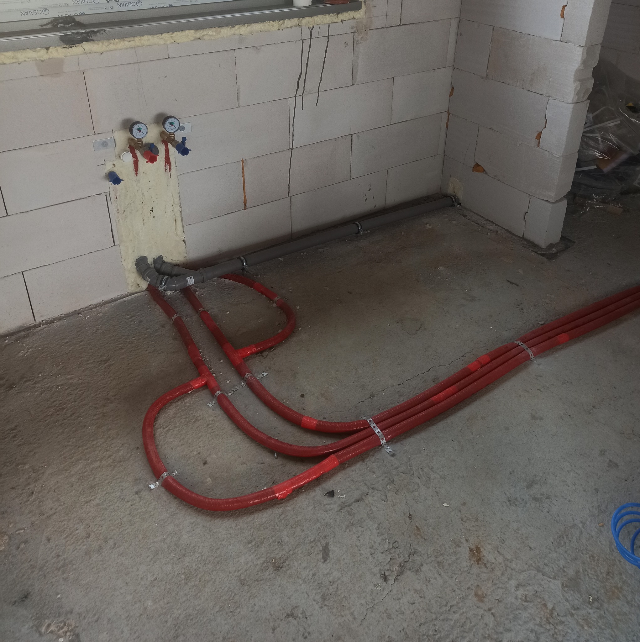 Montaż ogrzewania podłogowego w surowym stanie, widoczne czerwone rury mocowane do podłoża za pomocą metalowych uchwytów, ściany z białego bloczka.