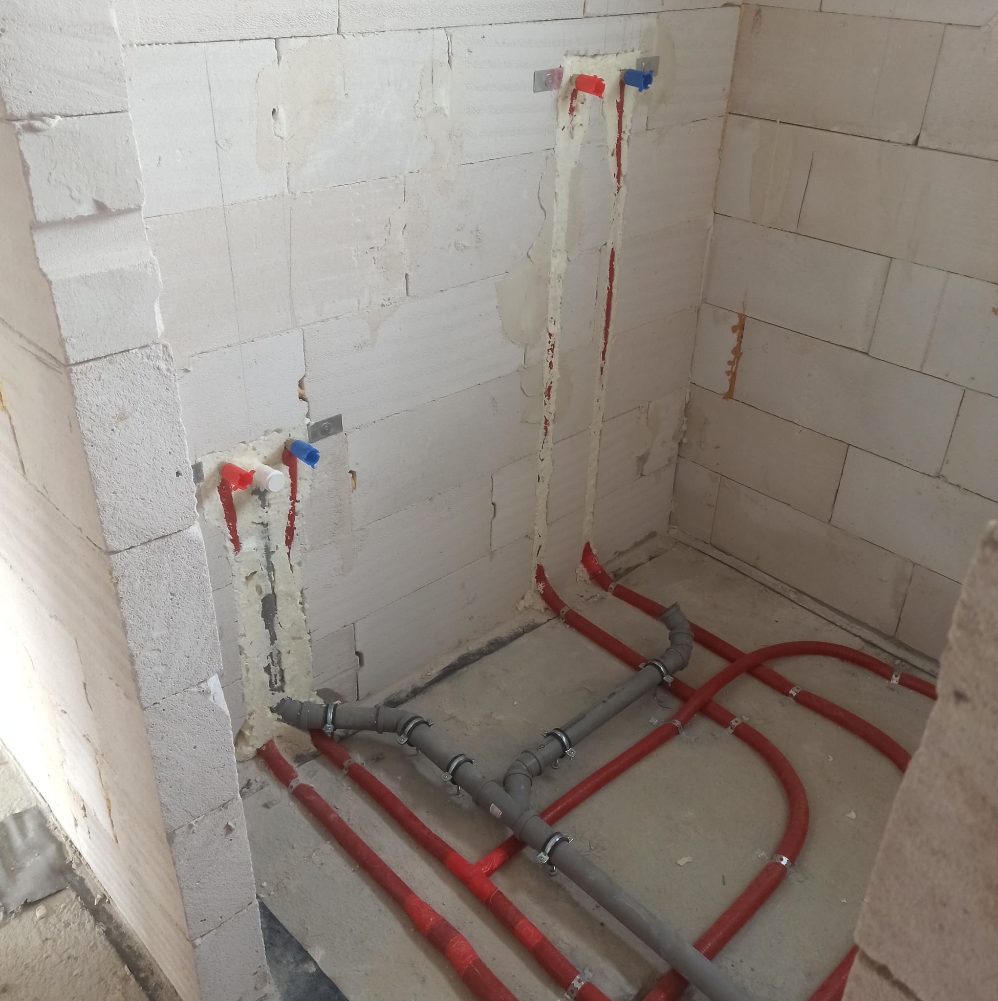 Wewnętrzna instalacja ogrzewania podłogowego w surowym stanie, widoczne rury grzewcze koloru czerwonego i szare rury kanalizacyjne z obejmami, ściany z białego betonu komórkowego.