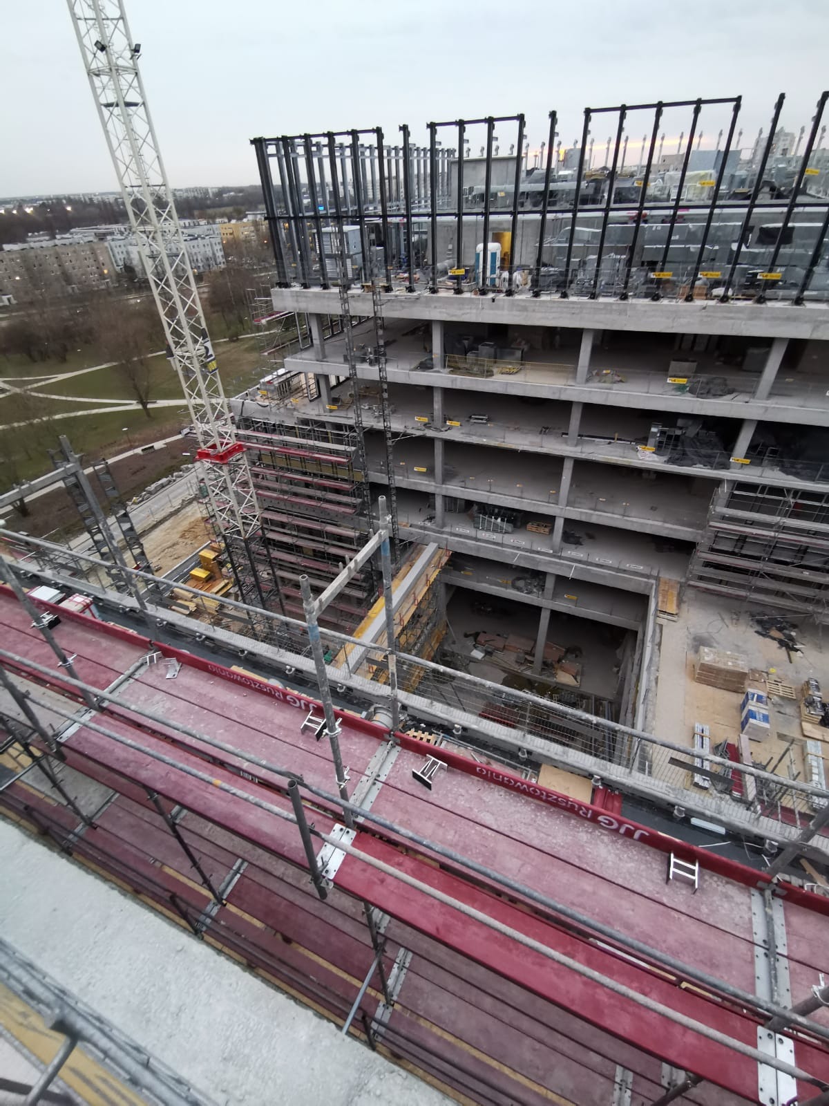 Budowa wielopiętrowego budynku w stanie surowym, widok z góry na rusztowania, dźwig i odsłonięte instalacje, w tle zabudowa miejska.