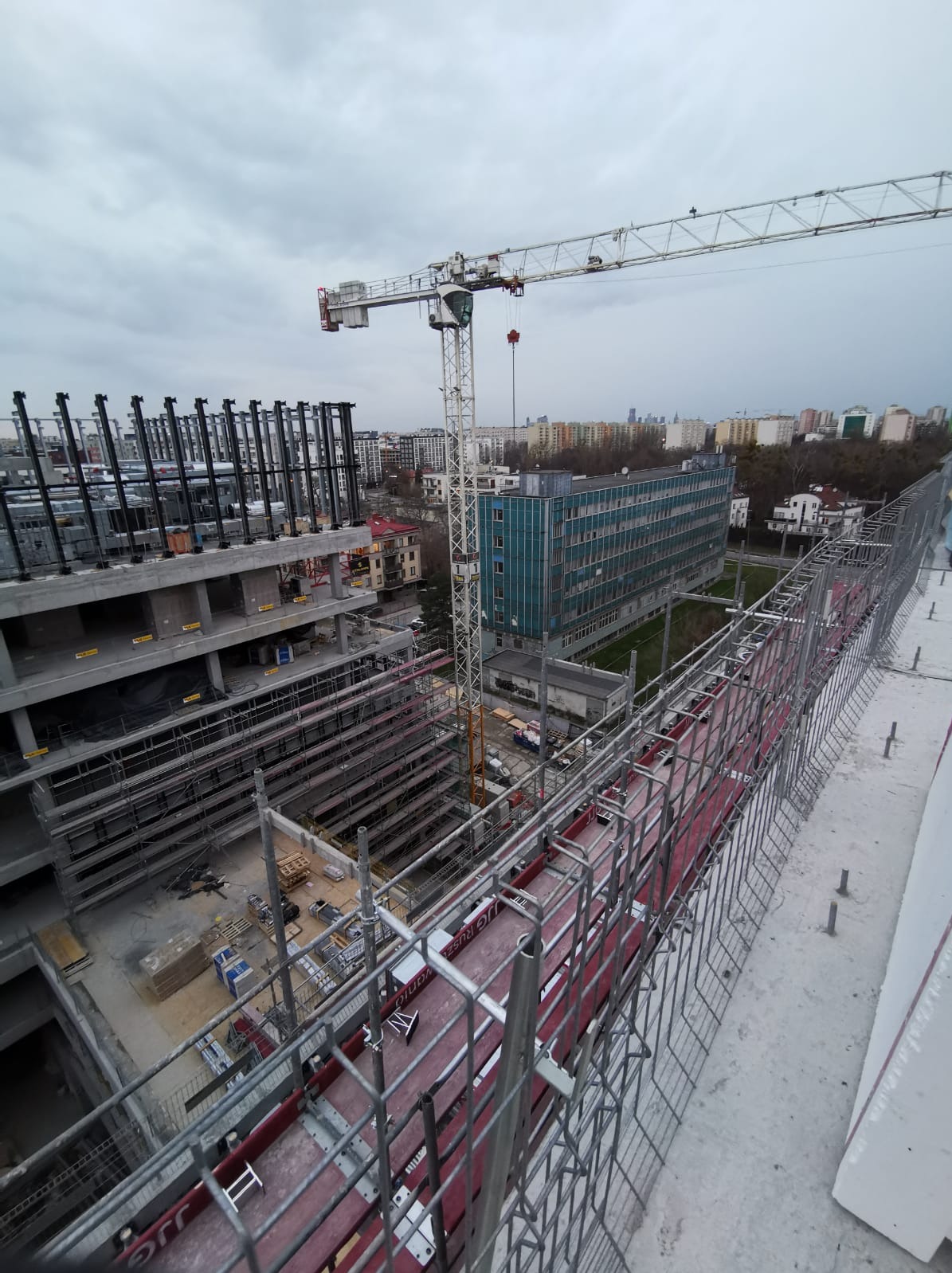 Widok z góry na plac budowy z rusztowaniami, żurawiem i formami szalunkowymi na krawędzi budynku, w tle zabudowa miejska i pochmurne niebo.