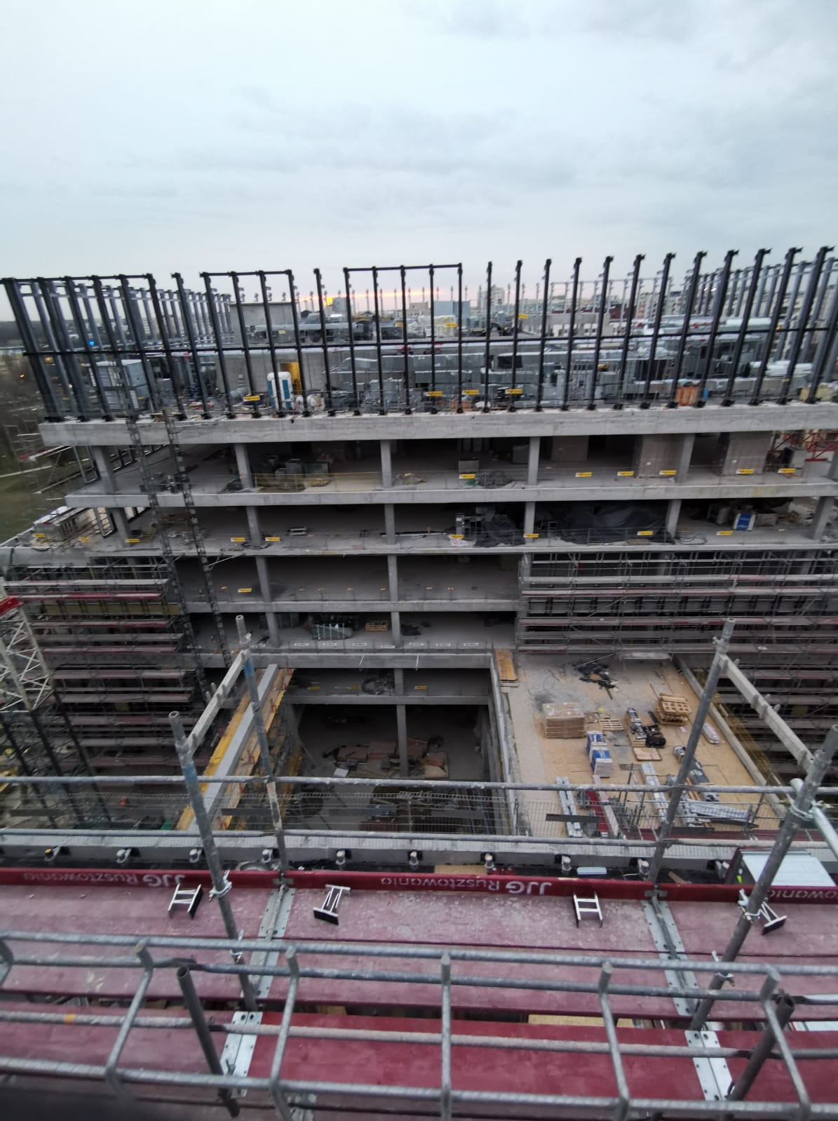 Wysoki budynek w stanie surowym z widocznymi instalacjami na dachu, sfotografowany z rusztowania w pochmurny dzień.