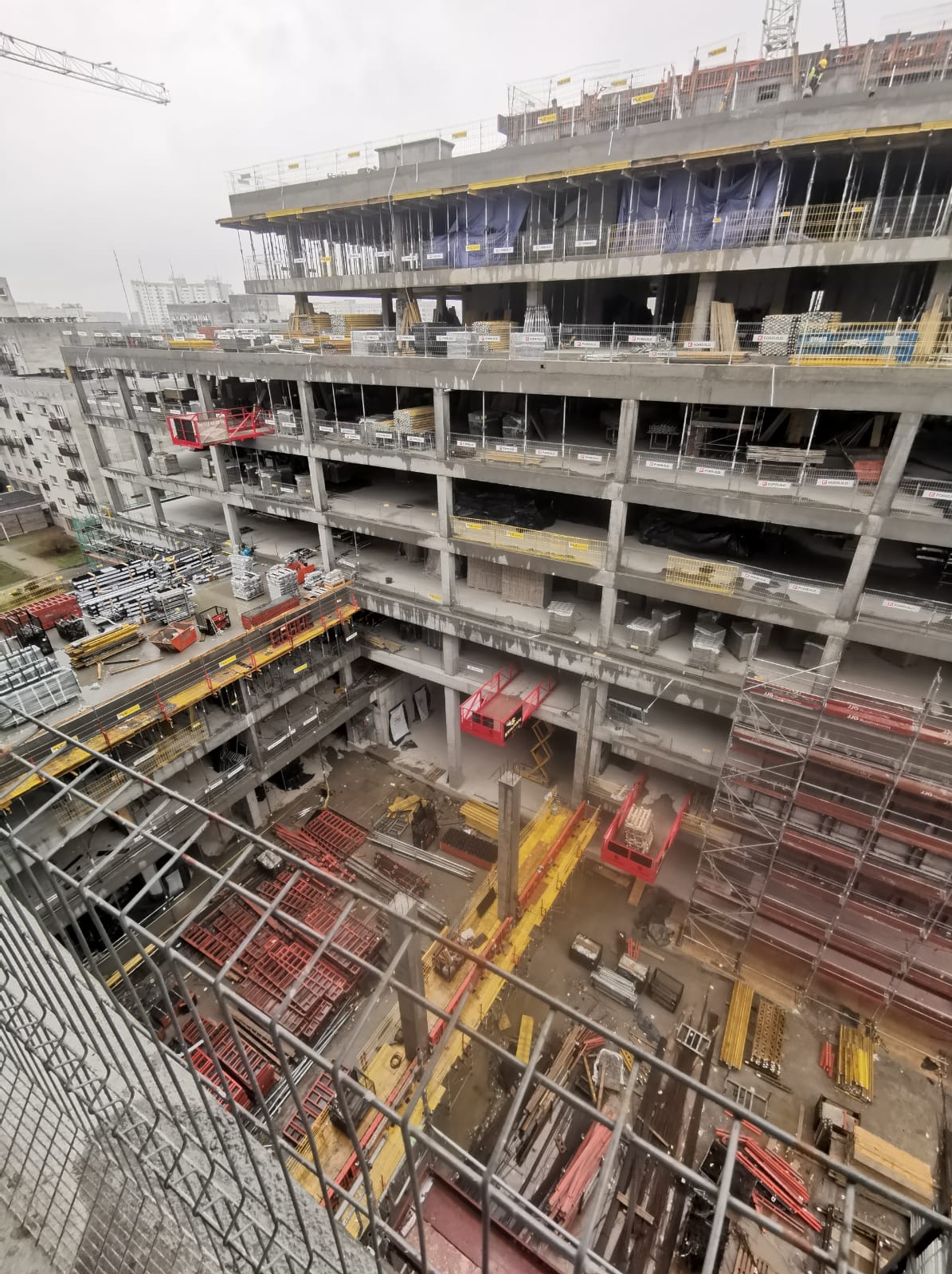 Wysoki budynek w stanie surowym, widok z góry na plac budowy z rusztowaniami, czerwonymi podnośnikami i materiałami budowlanymi, w tle zabudowa miejska w pochmurny dzień.