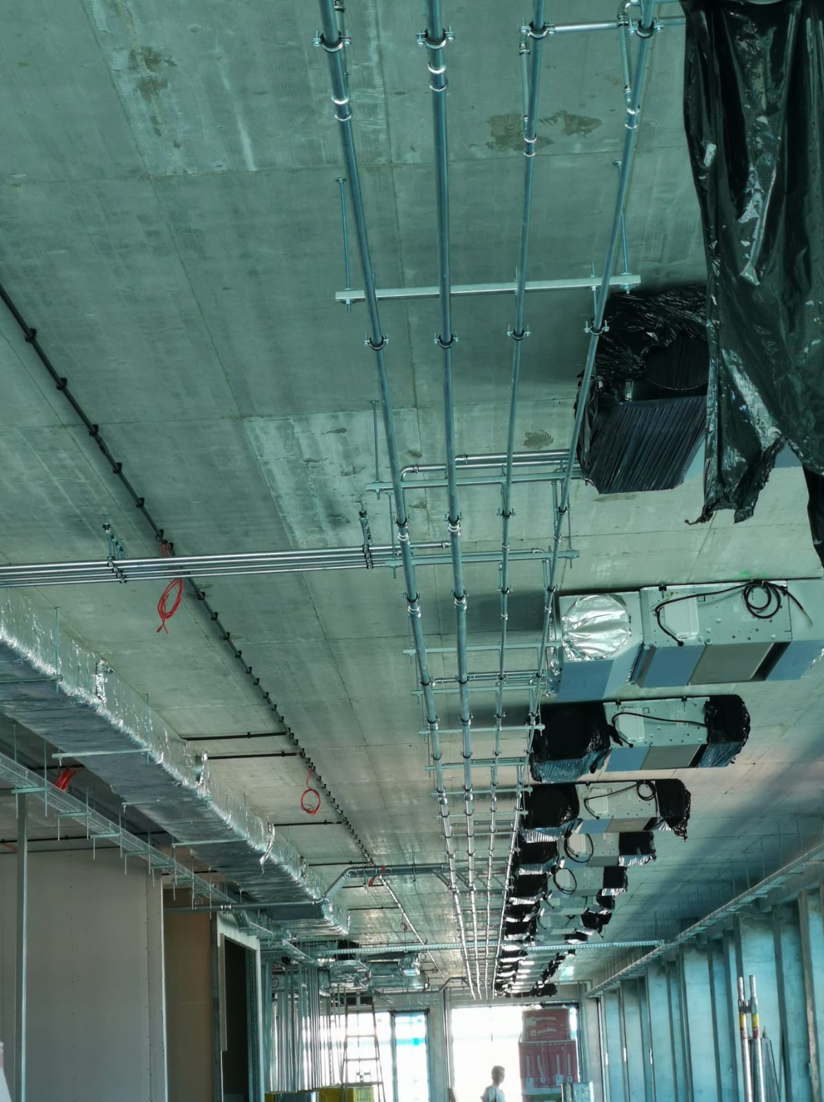 Instalacja rur i przewodów wentylacyjnych pod betonowym sufitem w budynku w trakcie budowy, widoczne elementy mocujące i fragmenty systemów wentylacyjnych.