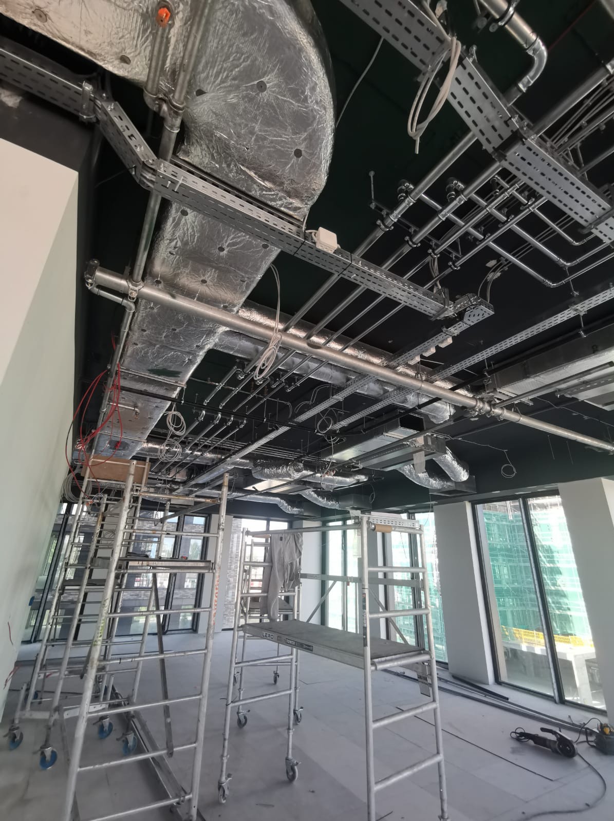 Widok instalacji pod sufitem w nowym biurze, rury w izolacji, koryta kablowe, drabiny rusztowaniowe w pomieszczeniu z dużymi oknami.