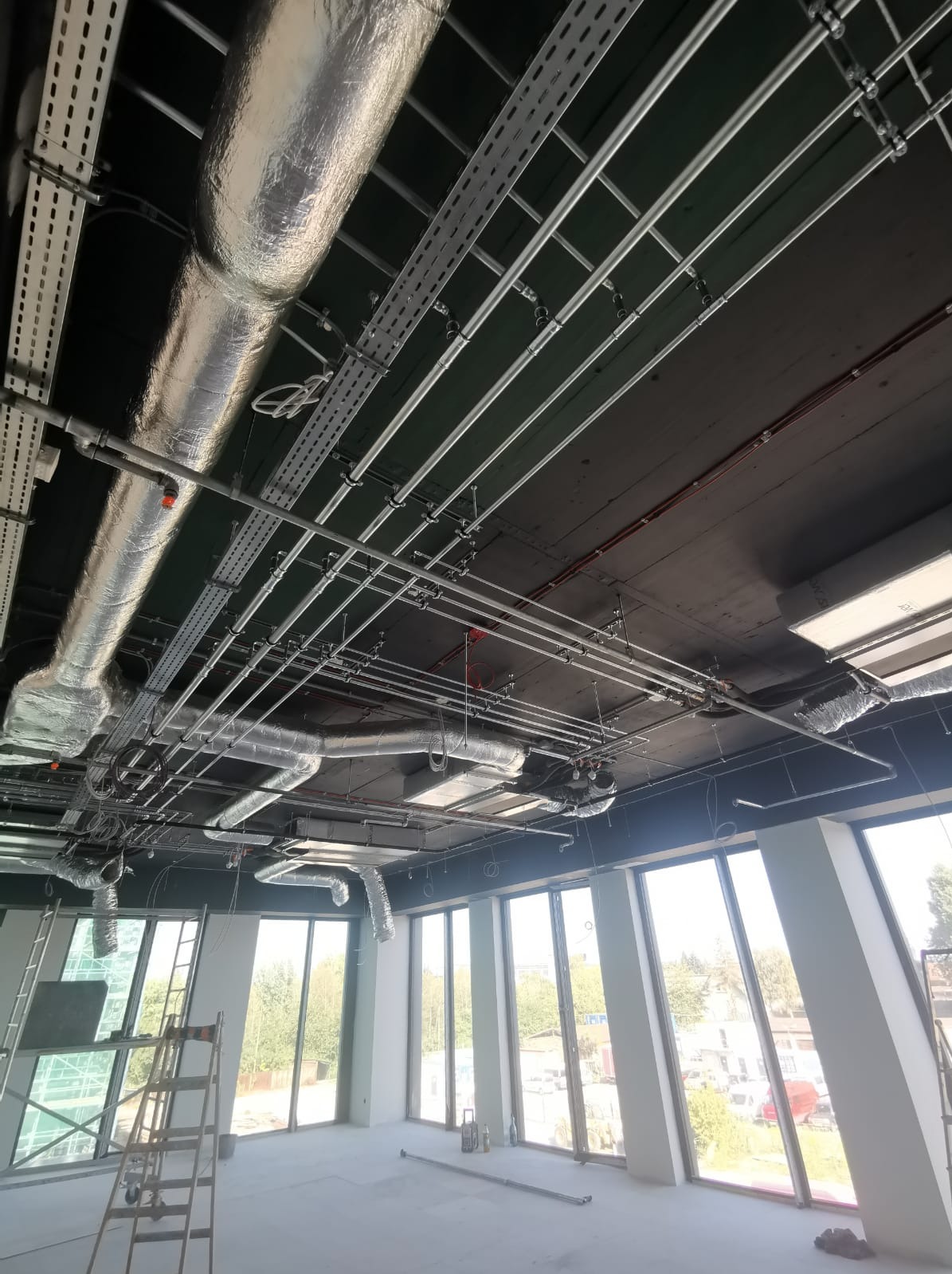 Widok z dołu na sufit w nowym budynku z widocznymi instalacjami: rurami wentylacyjnymi w srebrnej izolacji, metalowymi korytkami kablowymi i rurami instalacji przeciwpożarowej, z oknami na zewnątrz.