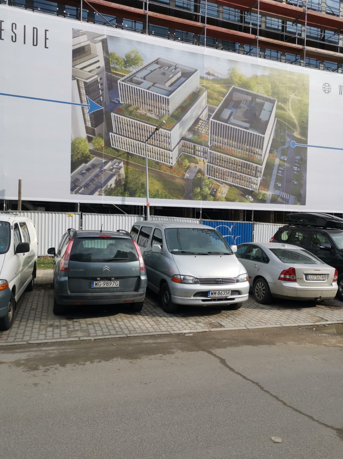 Widok na plac budowy z zaparkowanymi samochodami na pierwszym planie i dużym banerem reklamowym przedstawiającym wizualizację nowoczesnego biurowca w tle.