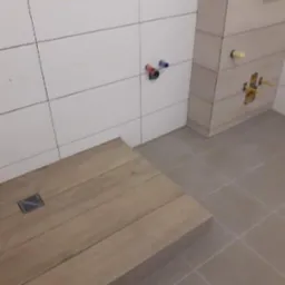 Płytki ceramiczne imitujące drewno na podłodze prysznica z odpływem liniowym, białe płytki ścienne z wyprowadzonymi instalacjami wodnymi.