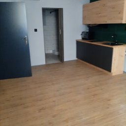 Ryszard Łaszewski - Wnętrze po remoncie: widok na nową podłogę z paneli, drzwi wejściowe, oraz zabudowę kuchenną z czarnymi frontami i drewnianymi szafkami wiszącymi.