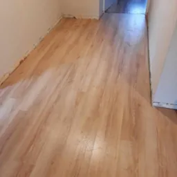 Nowo położone panele podłogowe w korytarzu, widoczne ślady po remoncie przy ścianach i otwarte drzwi w tle.